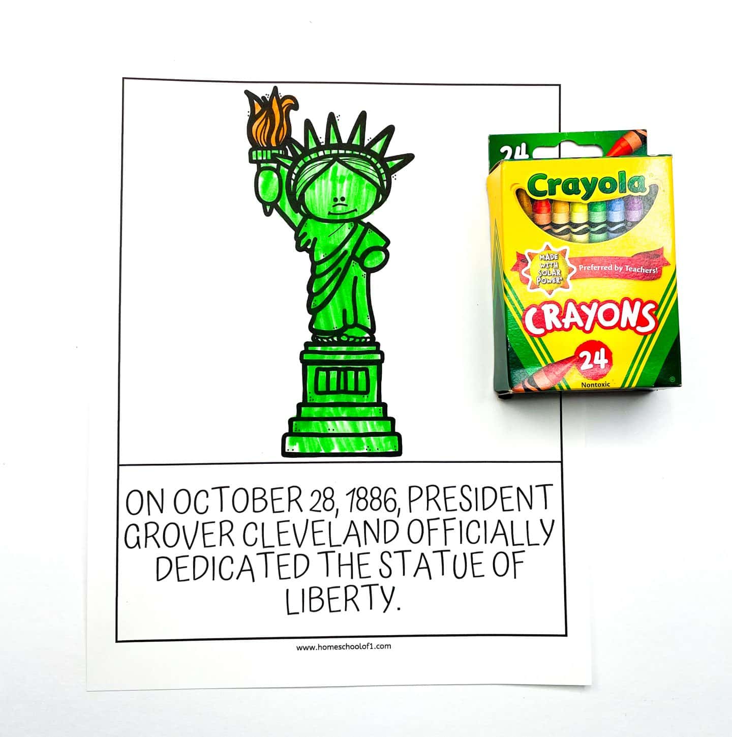 A kids’ coloring worksheet featuring a cartoon-style Statue of Liberty colored in green and orange. Below the image, text reads: “On October 28, 1886, President Grover Cleveland officially dedicated the Statue of Liberty.” A 24-pack of Crayola crayons is placed to the right of the worksheet on a white background.