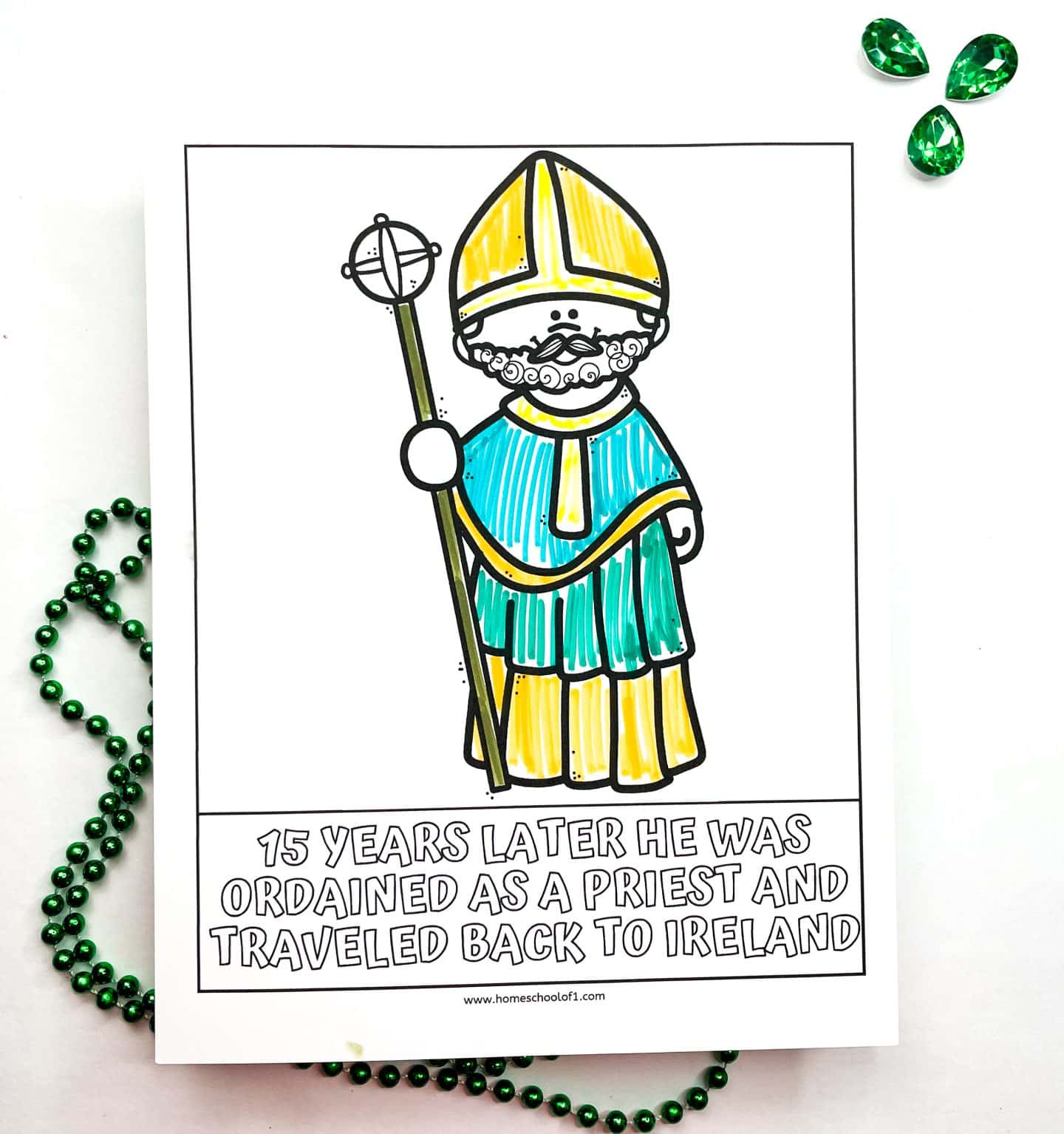 A St. Patrick’s Day worksheet featuring a hand-drawn, partially colored illustration of Saint Patrick in green and yellow robes holding a bishop's staff. The caption reads, “15 years later he was ordained as a priest and traveled back to Ireland.” Green bead garland and three green gem decorations are placed beside the worksheet.

