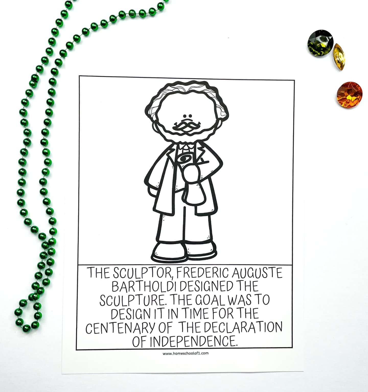 A black and white coloring worksheet featuring a cartoon image of sculptor Frédéric Auguste Bartholdi holding a small model of the Statue of Liberty. Below the illustration, the text reads: “The sculptor, Frederic Auguste Bartholdi designed the sculpture. The goal was to design it in time for the centenary of the Declaration of Independence.” The sheet is placed on a white background with a green beaded necklace and three colorful plastic gems.