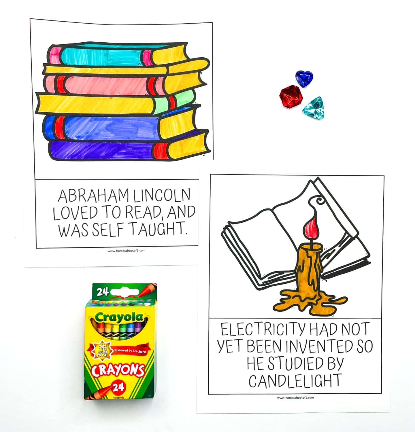 Two Abraham Lincoln-themed worksheets for kids, featuring colorful drawings of a stack of books and a candlelit book. One worksheet says “Abraham Lincoln loved to read, and was self taught,” while the other explains he studied by candlelight because electricity hadn't been invented yet. A box of Crayola crayons and three plastic gem shapes (red, blue, and teal) are also included in the flat lay.