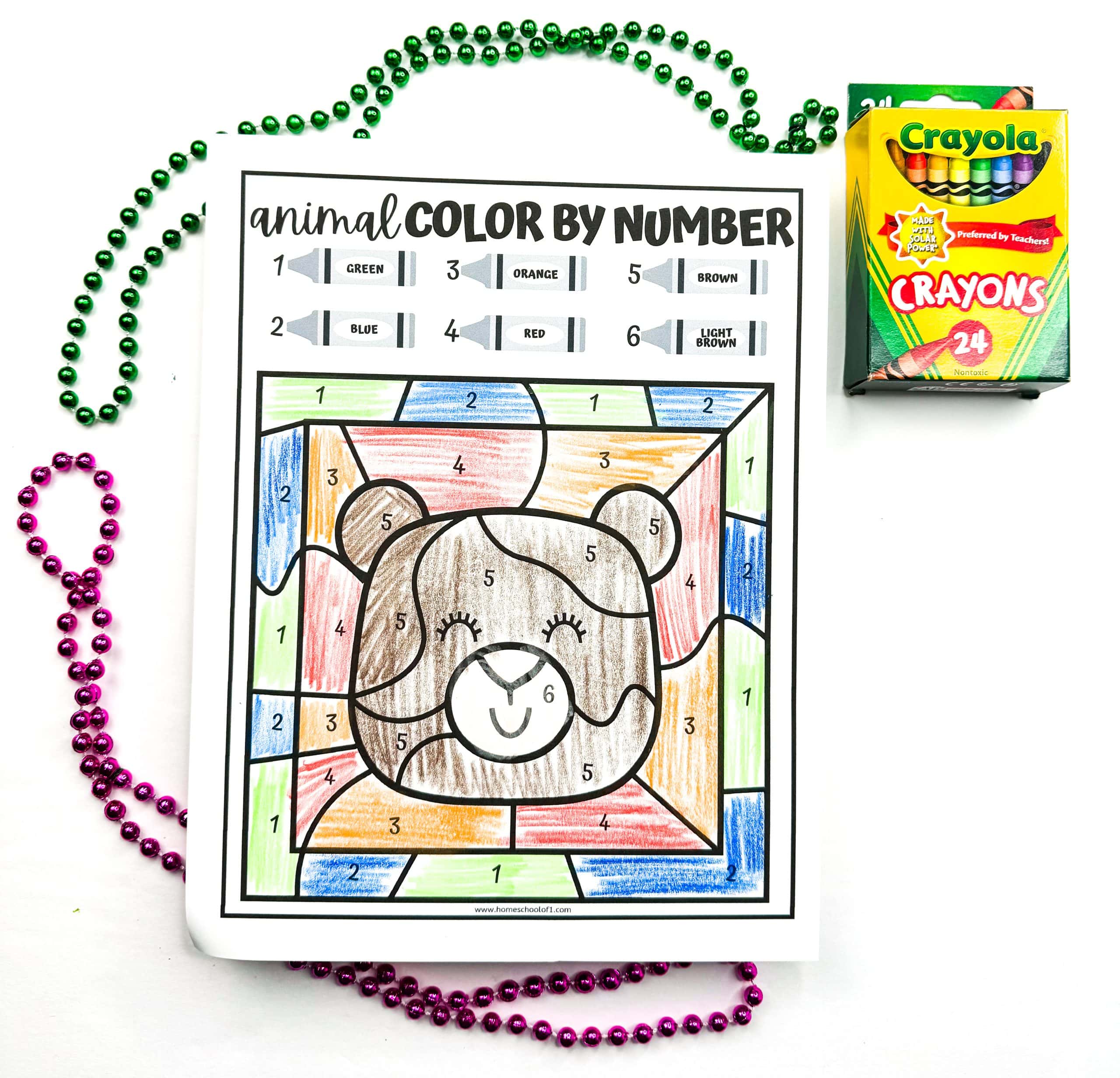 A completed bear color by number worksheet showing a cartoon bear face colored with crayons in brown, red, orange, green, blue, red, and light brown, matching the number key at the top. The worksheet is surrounded by green and pink beads, with a 24-pack of Crayola crayons in the upper right corner.