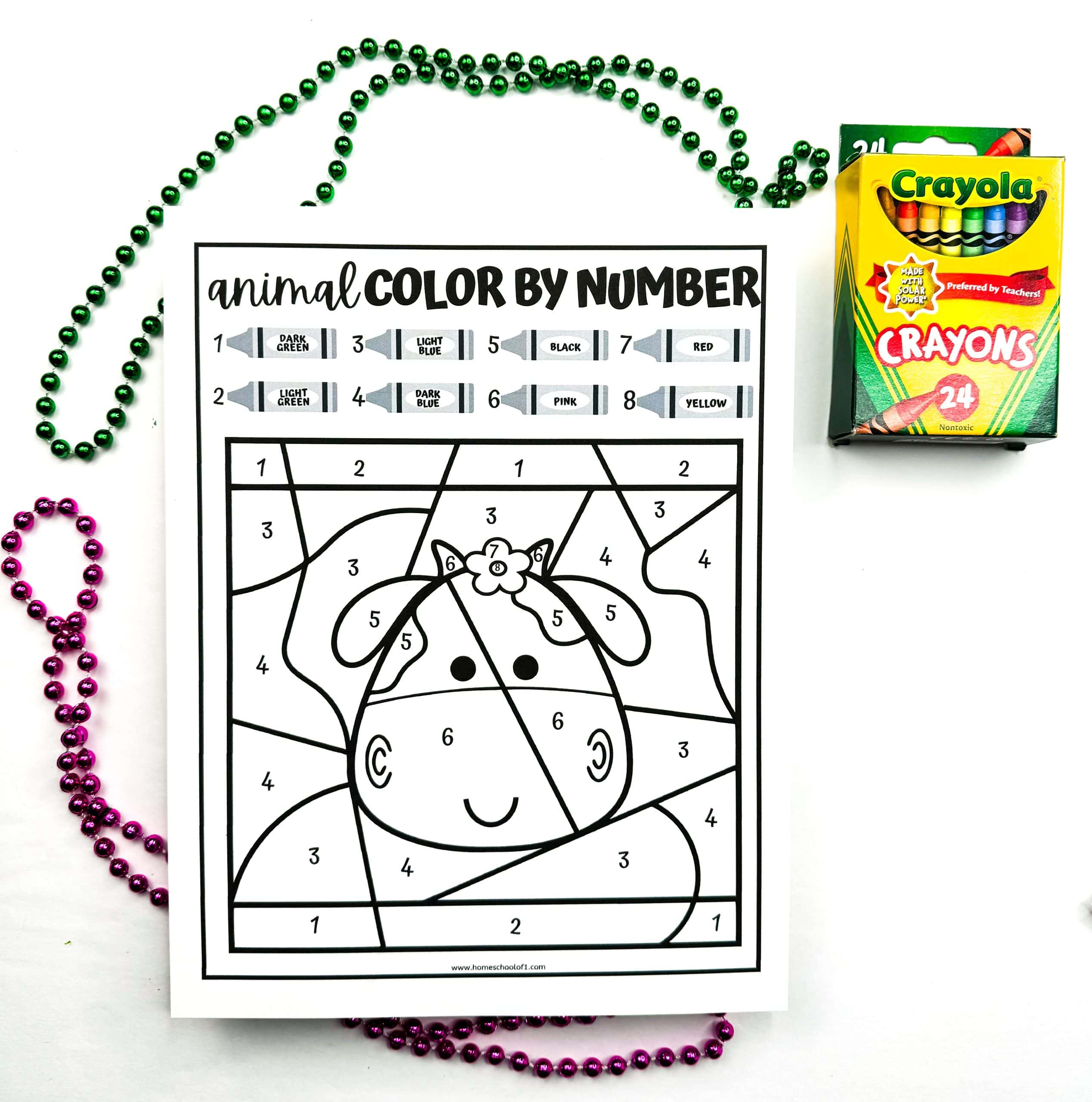 An animal color by number worksheet featuring a cartoon-style cow with a flower on its head. The uncolored image includes numbered sections with a crayon color key above listing dark green, light green, light blue, dark blue, black, pink, red, and yellow. The worksheet is placed beside a 24-pack of Crayola crayons and is surrounded by green and purple bead necklaces.