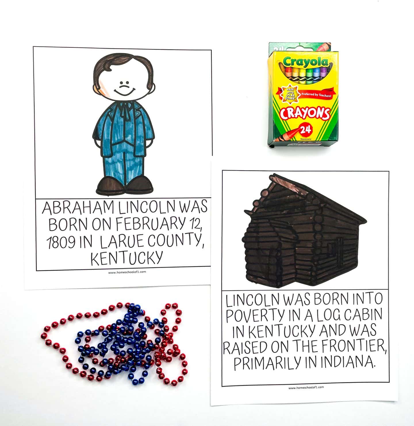 Flat lay of two illustrated Abraham Lincoln worksheets for kids, featuring a colored-in cartoon of Lincoln and a log cabin. One worksheet states Lincoln was born on February 12, 1809 in LaRue County, Kentucky; the other notes he was born into poverty and raised on the frontier. Red and blue beaded necklaces and a 24-pack of Crayola crayons are also pictured for a colorful, hands-on learning setup.