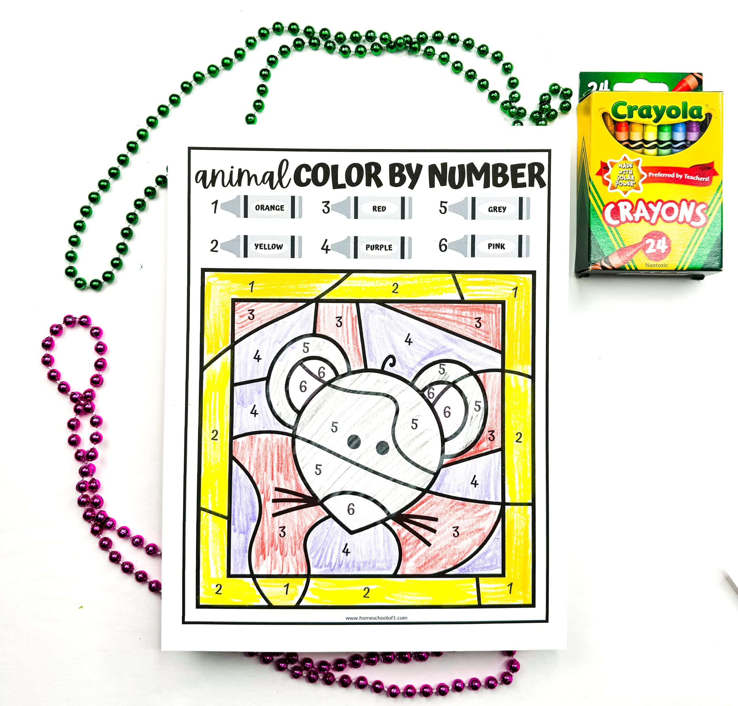 A finished animal color by number worksheet featuring a cartoon mouse, colored with crayons in orange, yellow, red, purple, grey, and pink according to the number guide at the top. The page is surrounded by green and purple bead necklaces and a box of 24 Crayola crayons is placed to the upper right.

