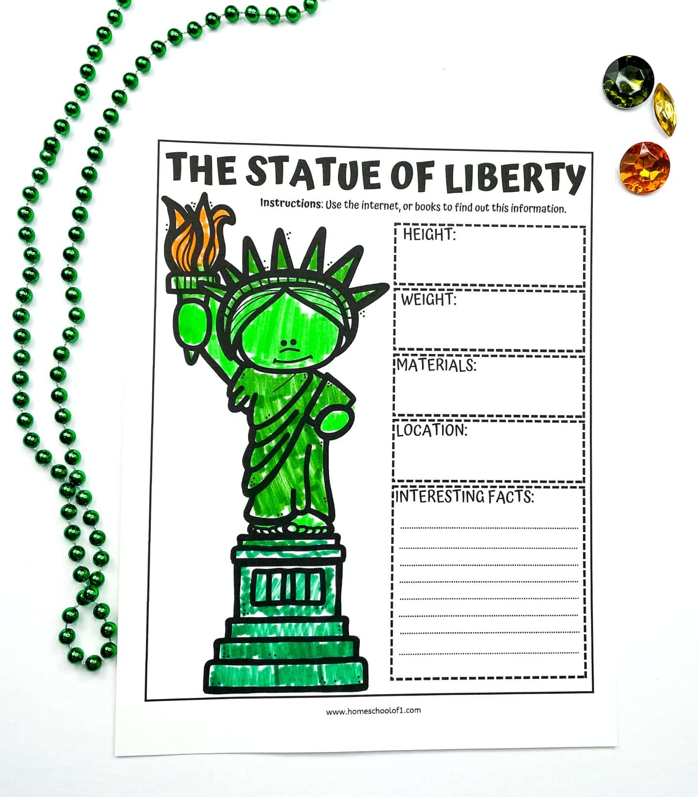  A Statue of Liberty worksheet for kids featuring a large, cartoon-style image of the statue colored in green and orange. The worksheet includes labeled boxes for students to fill in information such as height, weight, materials, location, and interesting facts. It's displayed on a white surface with a green beaded necklace and colorful plastic gems for decoration.