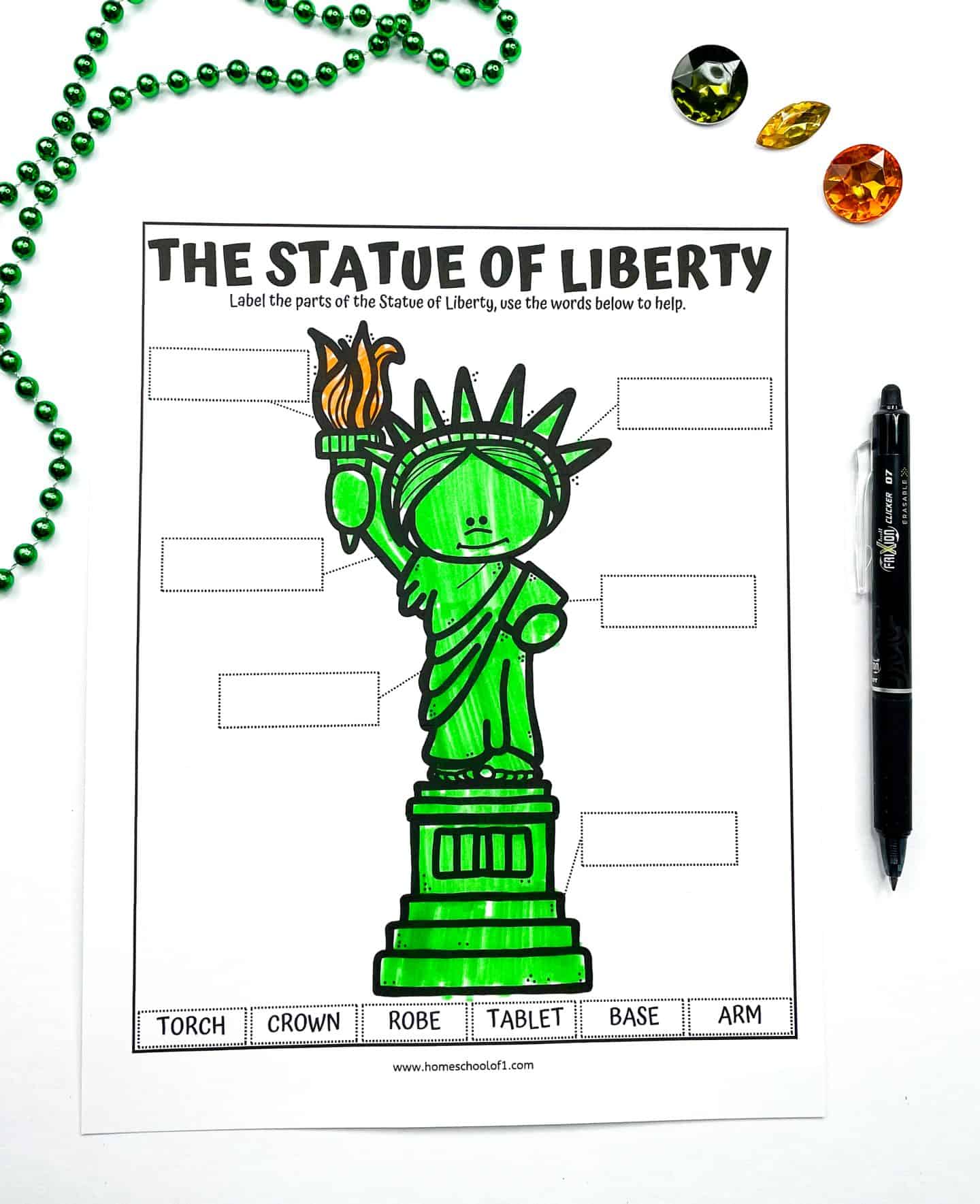 A kids' worksheet titled “The Statue of Liberty” with a large, colored illustration of the statue and blank labels for identifying its parts. Words at the bottom—torch, crown, robe, tablet, base, and arm—are provided to match with the correct parts. The page is displayed with a green beaded necklace, a black pen, and three colorful plastic gems on a white background.