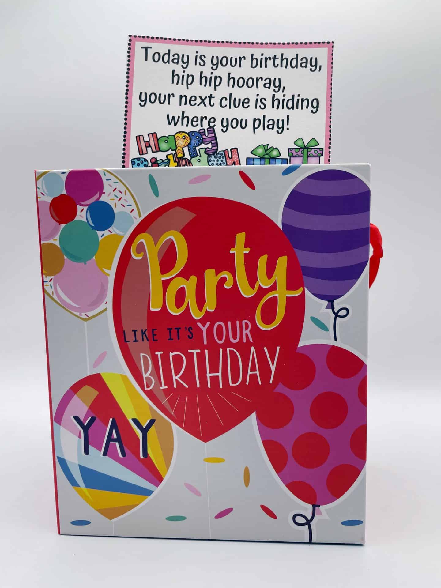 A festive gift bag decorated with colorful balloons and the phrase “Party like it’s your birthday” with a birthday scavenger hunt clue card sticking out of the top. The clue reads, “Today is your birthday, hip hip hooray, your next clue is hiding where you play!” making it a playful and fun start to a birthday scavenger hunt for kids.