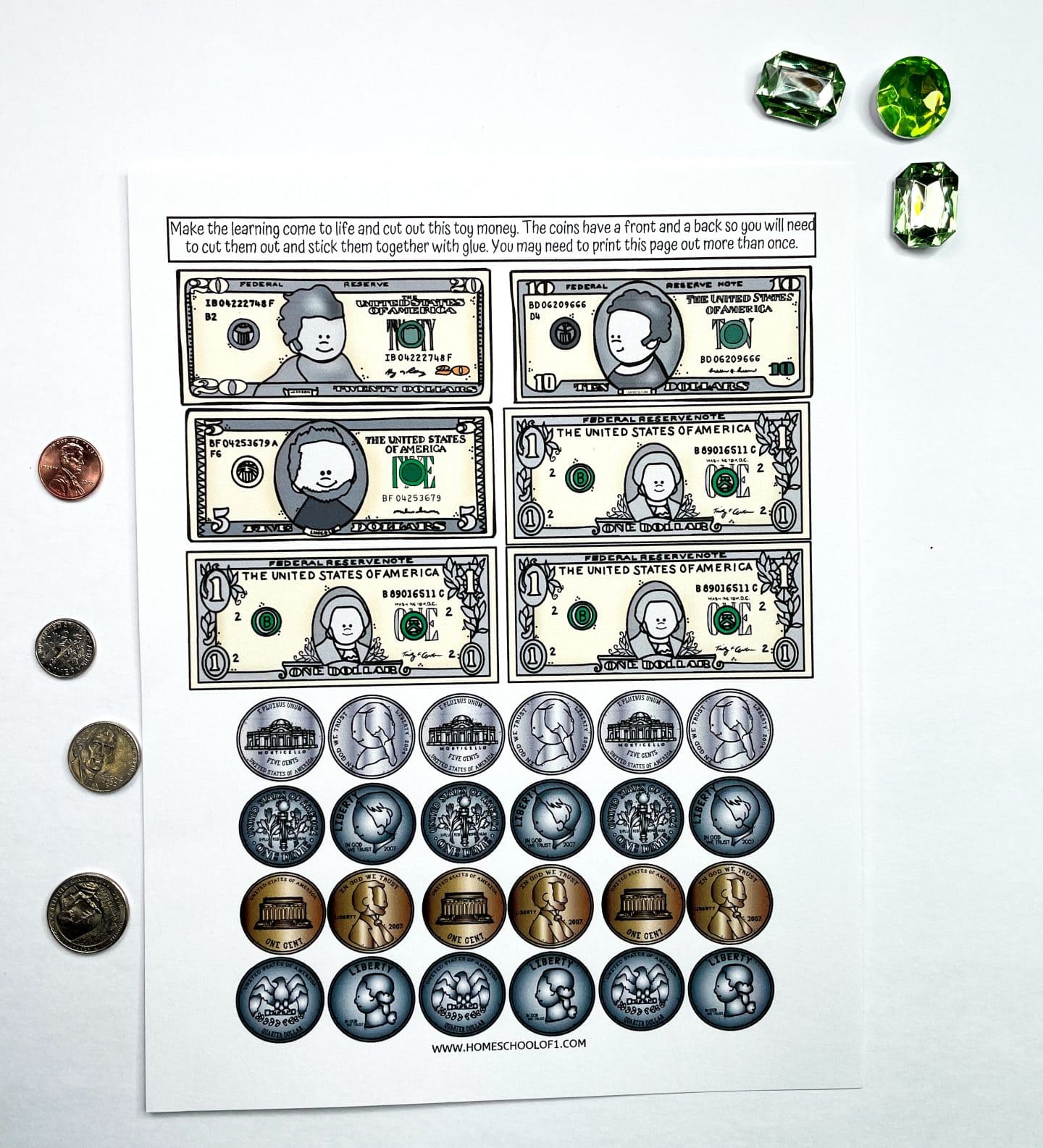 Printable play money sheet with illustrated U.S. currency, including $1, $5, and $10 bills and various coins like pennies, nickels, dimes, and quarters. Real coins are placed beside the sheet for comparison, and green gem counters add a playful touch.