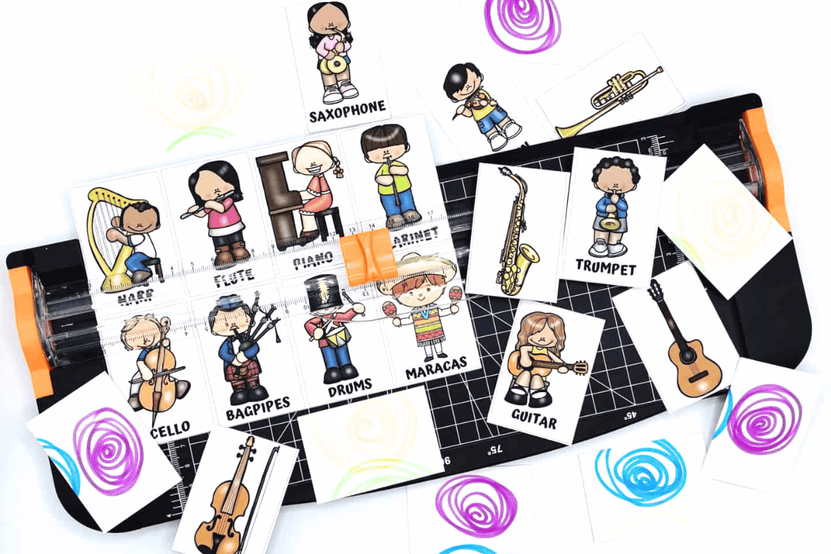 Close-up of a printable music-themed matching game in progress, with illustrated cards showing children playing instruments like harp, flute, piano, clarinet, cello, bagpipes, drums, maracas, saxophone, trumpet, and guitar. Some cards display instrument names and characters, while others are flipped to show colorful swirl patterns. A paper trimmer and scattered cards suggest the game is being prepped or played.