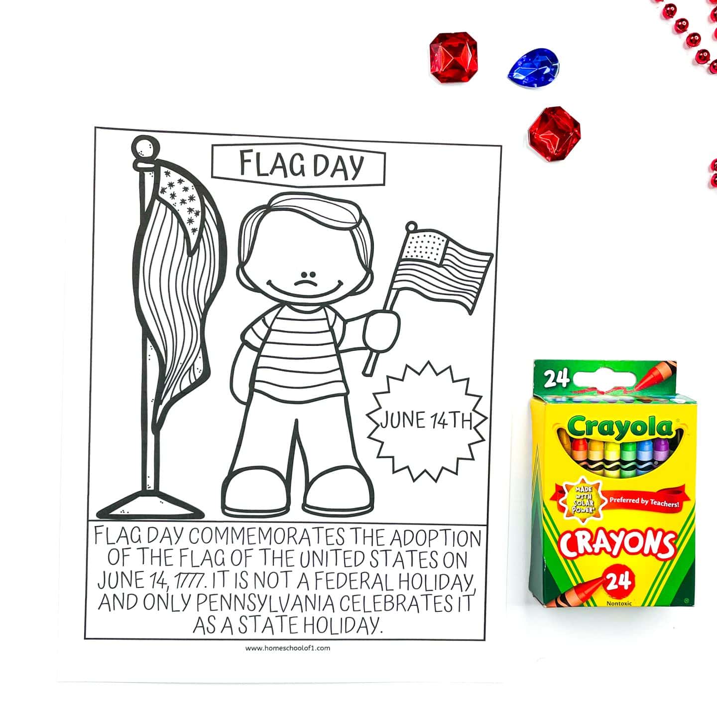 A Flag Day coloring page featuring a child holding a small U.S. flag next to a larger American flag on a flagpole. The page includes the date “June 14th” and a caption that explains Flag Day commemorates the adoption of the U.S. flag on June 14, 1777. Red and blue gem decorations and a 24-pack of Crayola crayons are placed beside the page.