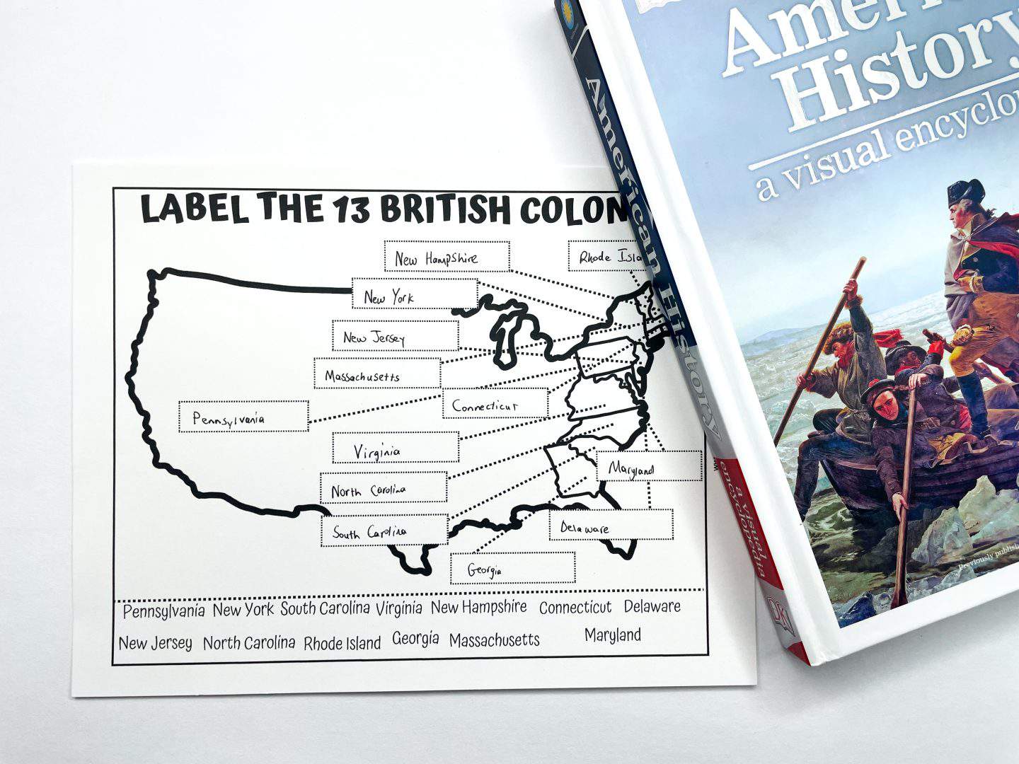 a "Label the 13 British Colonies" worksheet with a map of the United States and labeled boxes for each of the original 13 colonies. A book titled "America’s History: A Visual Encyclopedia" and a black pen are visible beside the worksheet.