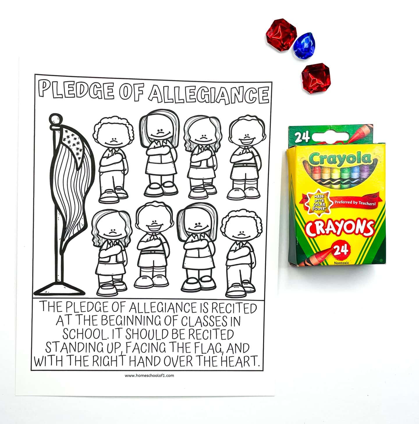Black-and-white Pledge of Allegiance coloring page showing a group of cartoon children standing by an American flag with their hands over their hearts. Below the image is the full instruction: “The Pledge of Allegiance is recited at the beginning of classes in school…” A box of Crayola crayons and three red and blue plastic gems are placed to the right of the sheet.