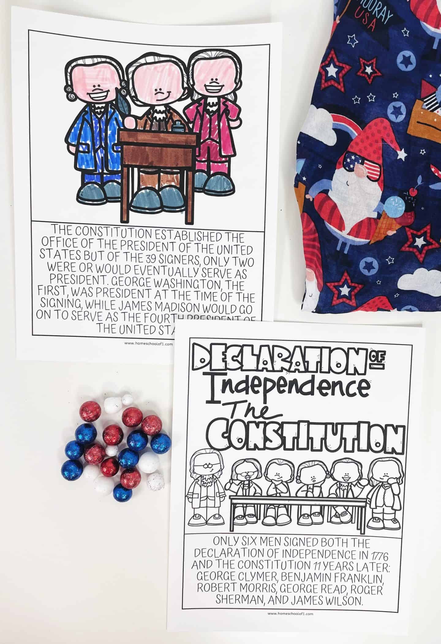 Two educational worksheets: one shows the three signers of the Constitution and mentions George Washington and James Madison becoming U.S. Presidents, with a description of their roles. The other features the six men who signed both the Declaration of Independence and the Constitution, including Benjamin Franklin. There are also red, white, and blue candies and a festive patterned fabric with stars and gnomes.