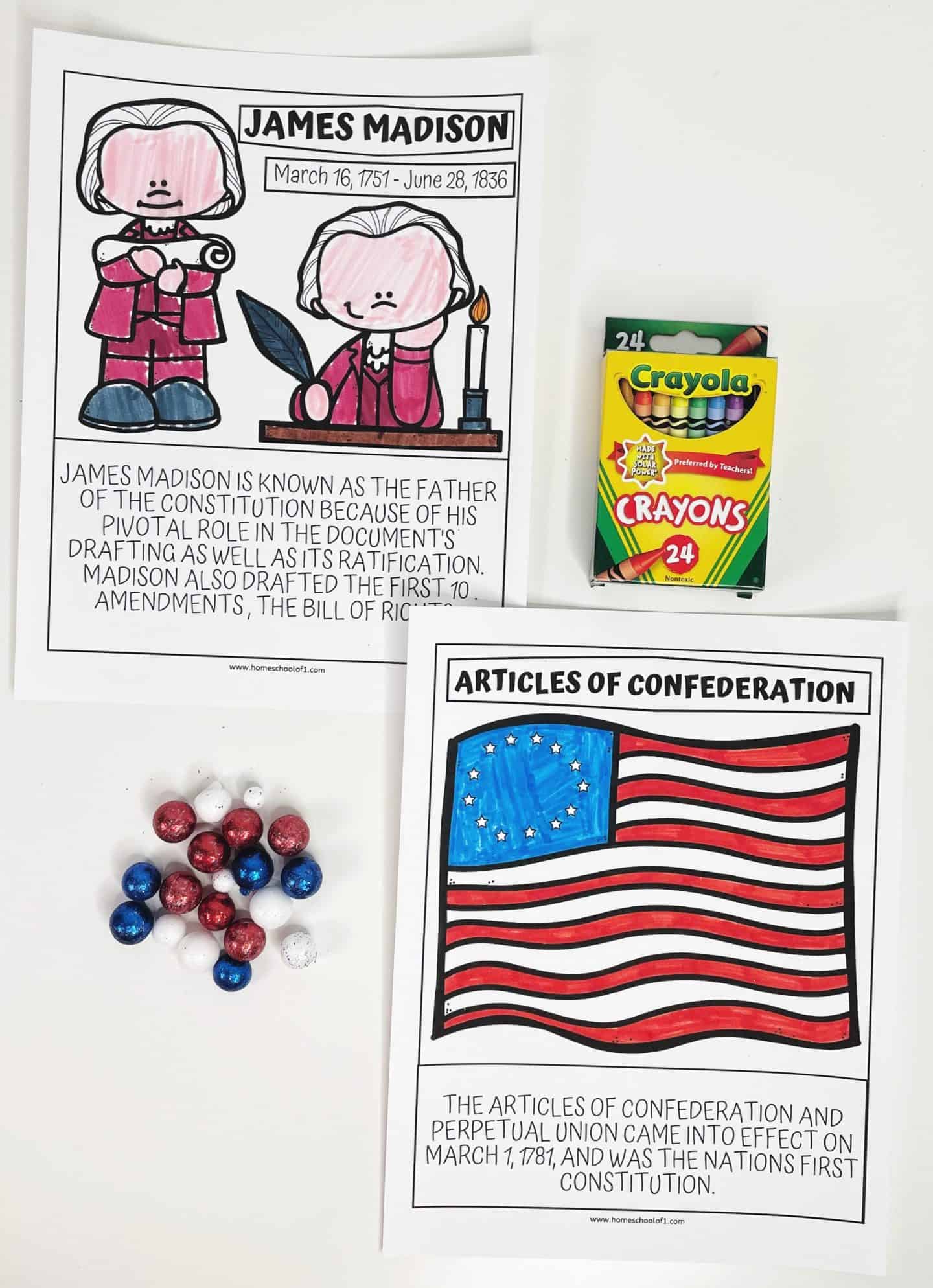 Two educational worksheets about James Madison and the Articles of Confederation. The James Madison worksheet includes a drawing of Madison with a description highlighting his role as the "Father of the Constitution." The Articles of Confederation worksheet features a drawing of the historical flag with a brief explanation about the Articles' implementation. Nearby are colorful candies in red, white, and blue, along with a box of Crayola crayons.
