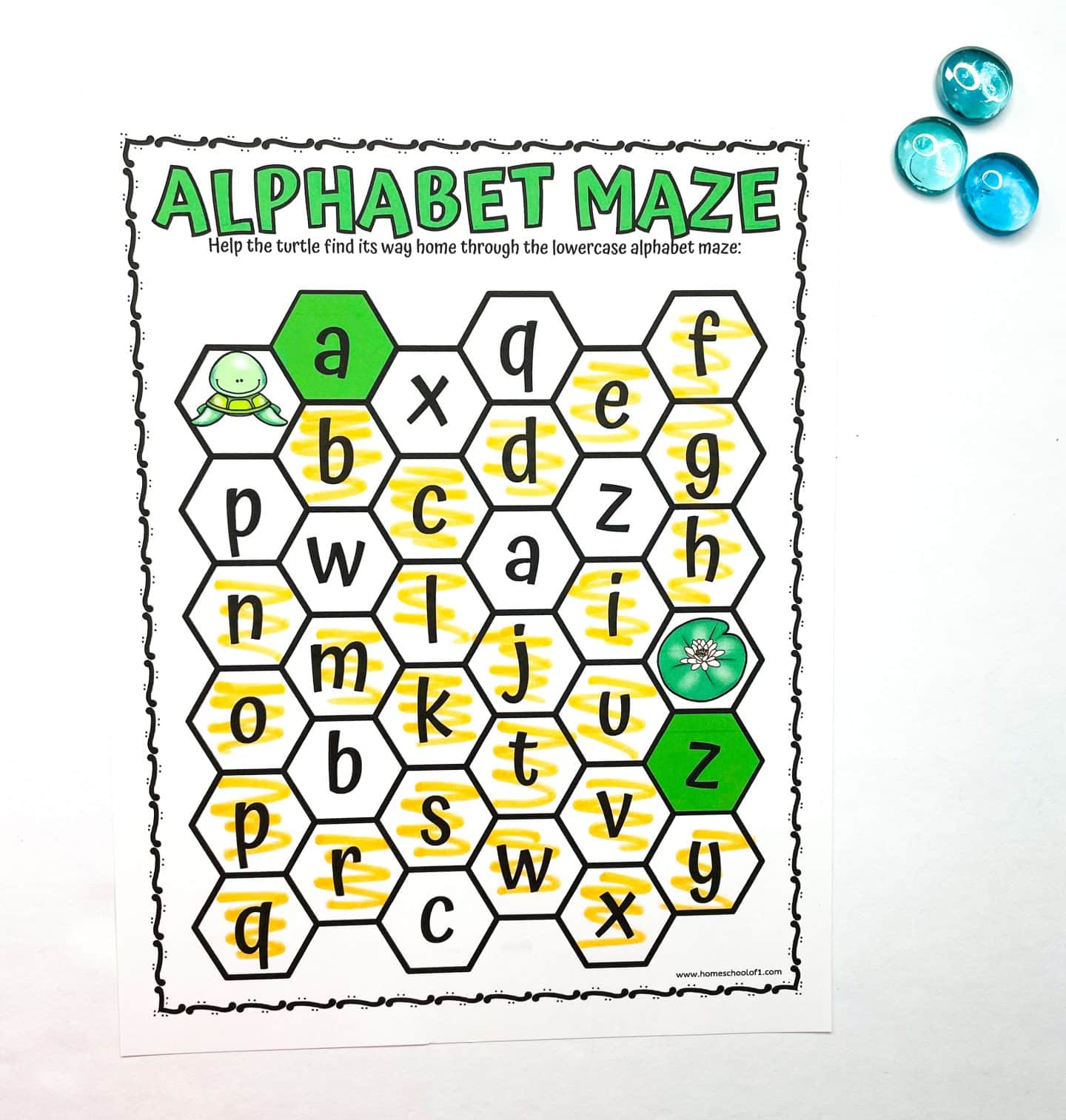 Preschool alphabet maze worksheet titled “Alphabet Maze” where kids help a turtle find its way home by following the lowercase alphabet in order. The maze consists of hexagons with letters, and the correct path from “a” to “z” is highlighted in yellow. A green turtle illustration marks the start, and a flower indicates the finish. Three blue counting gems are placed beside the worksheet.
