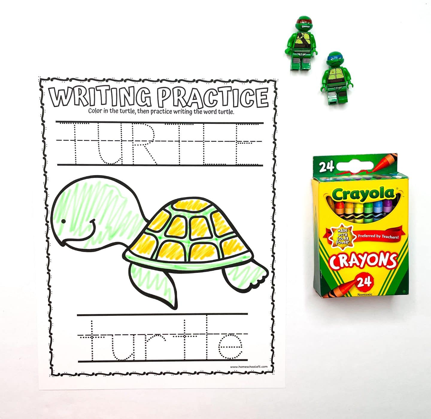 Preschool writing practice worksheet featuring the word "TURTLE" in uppercase and lowercase dotted lines for tracing. A cute turtle illustration is partially colored in green and yellow. To the side are a box of Crayola 24-count crayons and two small turtle-themed LEGO minifigures.