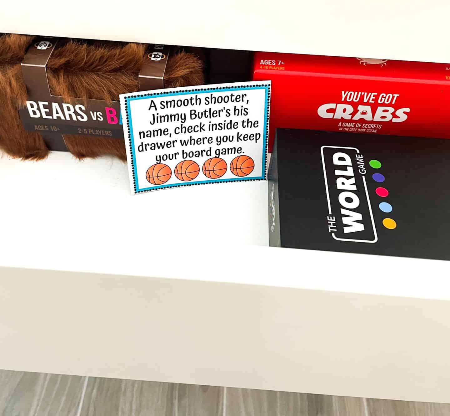 A drawer containing board games such as Bears vs Babies, You’ve Got Crabs, and The World Game. Inside the drawer is a basketball-themed scavenger hunt clue card that reads: “A smooth shooter, Jimmy Butler’s his name, check inside the drawer where you keep your board game,” decorated with four orange basketball icons.