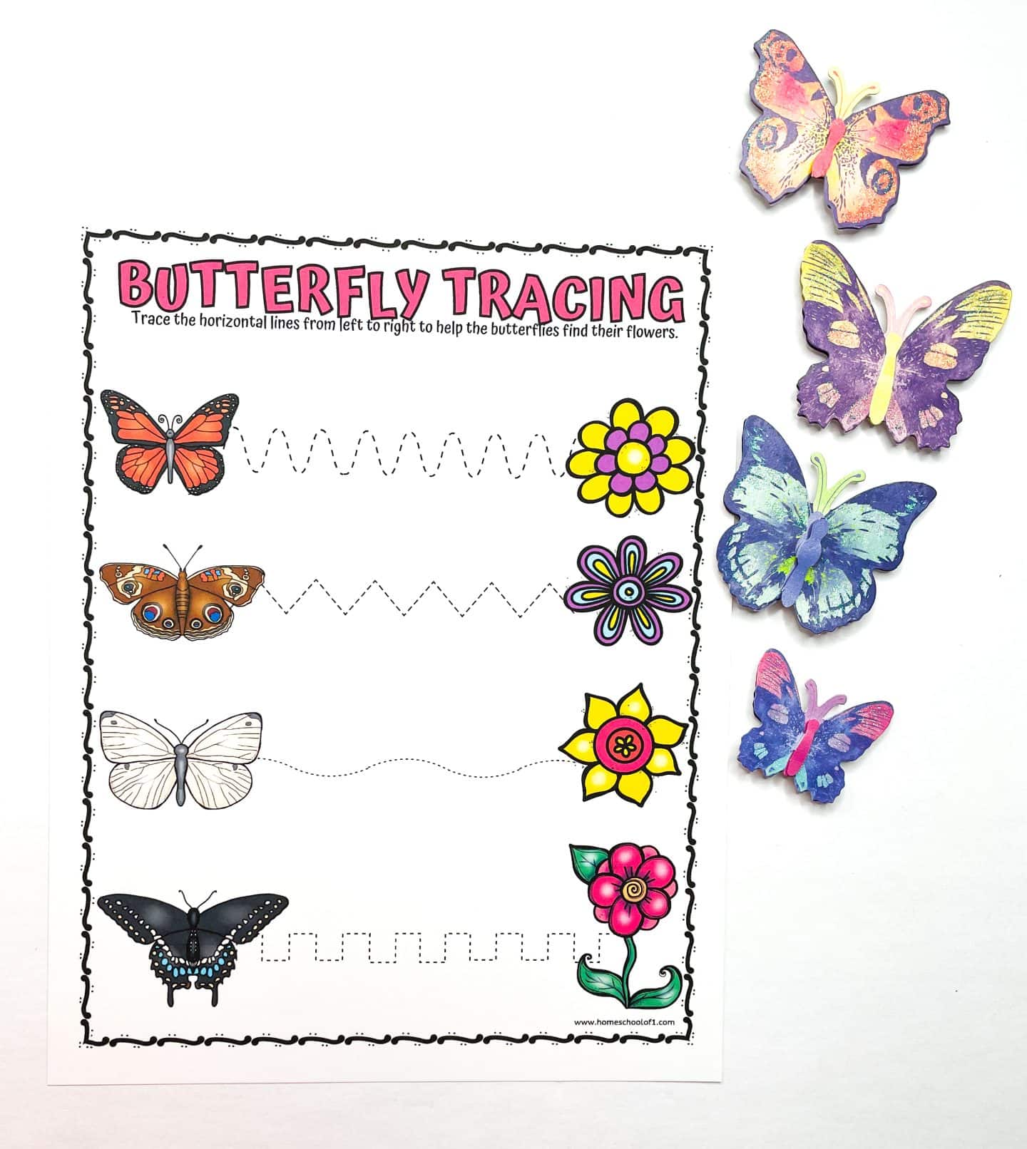 Butterfly prewriting practice worksheet featuring four black-and-white butterflies on the left and colorful flowers on the right, connected by different traceable lines to improve fine motor skills. Ideal for preschoolers learning pencil control and pattern tracing. Crayola crayons and paper butterfly cutouts decorate the scene.