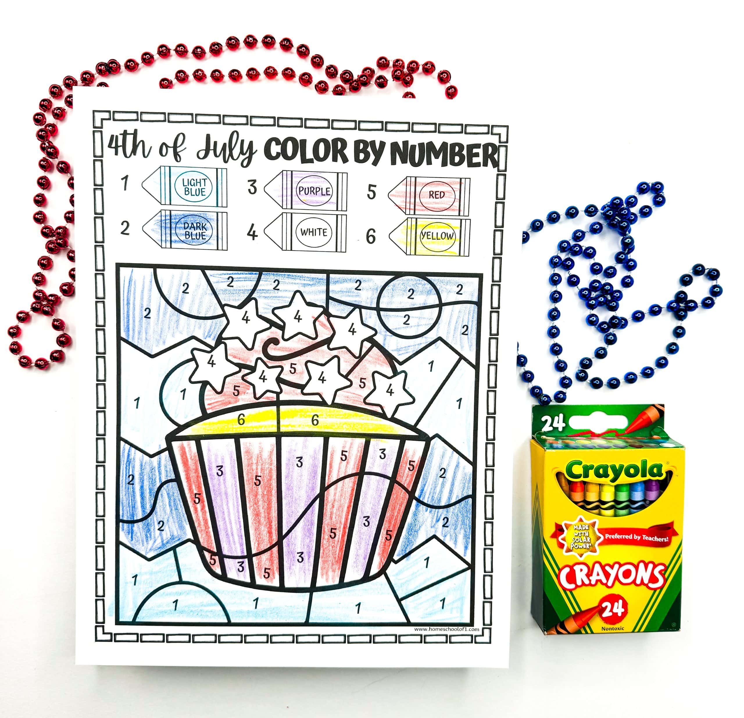 A completed 4th of July color-by-number worksheet features a festive cupcake with stars on top, decorated using light blue, dark blue, purple, red, white, and yellow based on the crayon key shown above. The coloring page is surrounded by red and blue bead necklaces and a 24-pack of Crayola crayons, enhancing the fun and patriotic celebration theme.