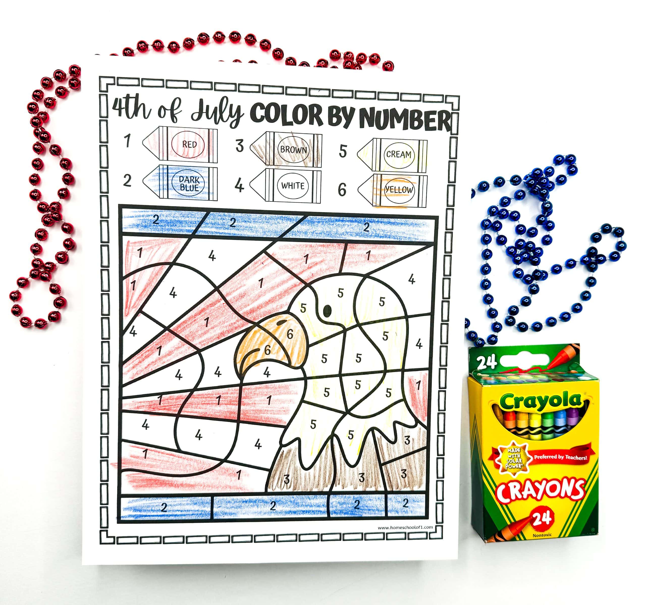 A completed 4th of July color-by-number worksheet showcases a bald eagle in front of a red and white striped background with a blue border. The eagle is colored using white, cream, yellow, and brown crayons, based on the color key at the top. Red and blue bead necklaces and a box of Crayola crayons frame the patriotic artwork