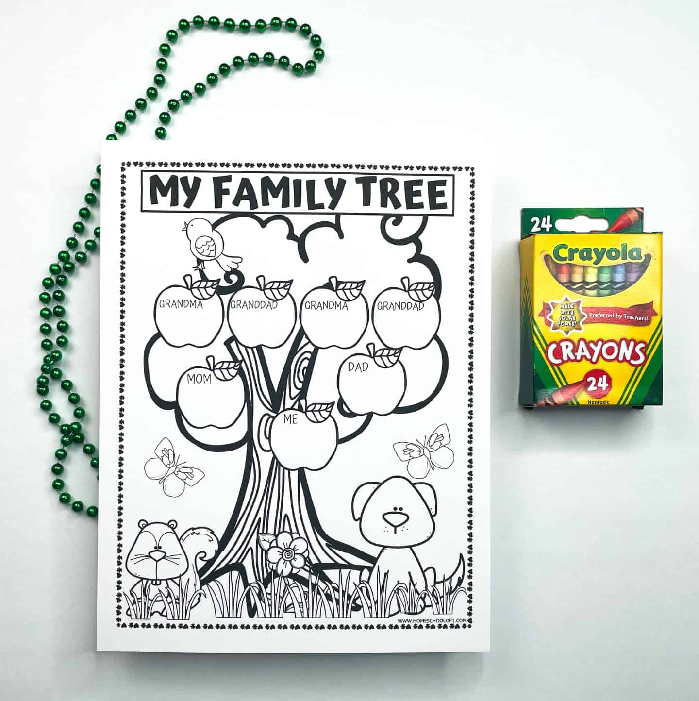 Black-and-white printable “My Family Tree” worksheet designed for kids, featuring a tree with apple-shaped spaces labeled for Grandma, Granddad, Mom, Dad, and Me. The base of the tree includes cute illustrations of a bear, a dog, butterflies, and flowers. A box of 24 Crayola crayons sits on the right, and a green beaded necklace is coiled on the left, suggesting the activity is ready to be colored.

