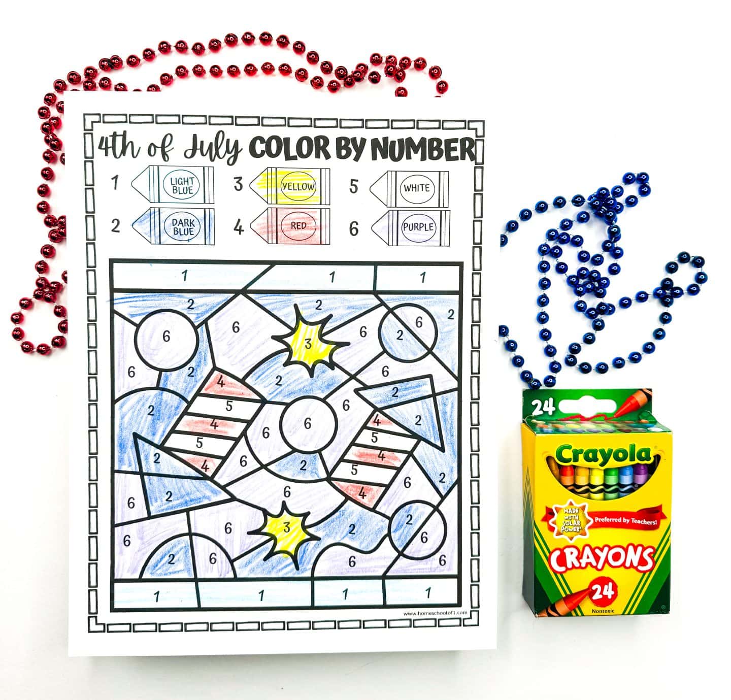 A completed 4th of July color-by-number worksheet displays two American flags and starburst shapes colored in light blue, dark blue, red, yellow, white, and purple according to the numbered crayon key at the top. The scene is surrounded by festive red and blue bead necklaces and a box of Crayola crayons, creating a cheerful Independence Day vibe.