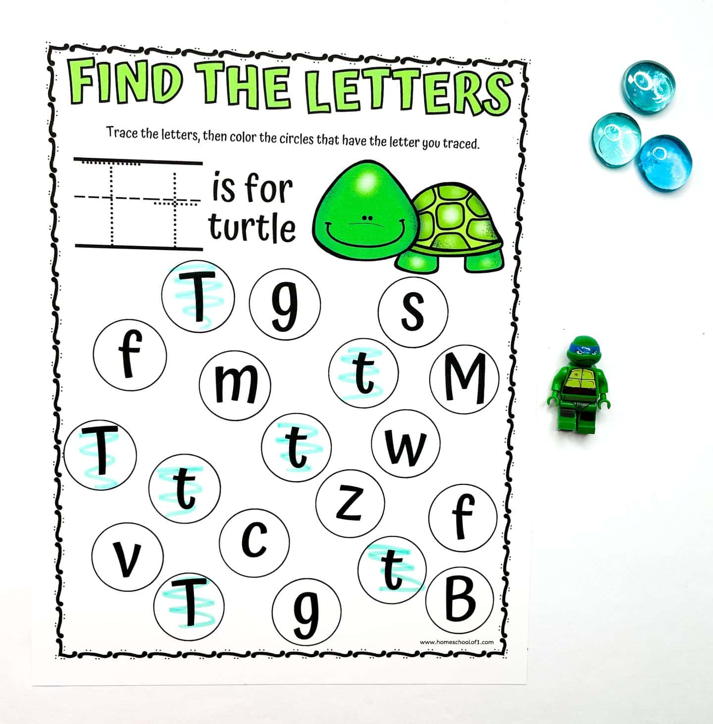 Preschool worksheet titled “Find the Letters” featuring a green turtle illustration and the prompt “T is for turtle.” Kids are instructed to trace the uppercase and lowercase “T” and then color the matching letters in the circle grid below. Several “T” and “t” letters have been highlighted in blue. A green turtle LEGO minifigure and three blue counting gems are placed beside the page.
