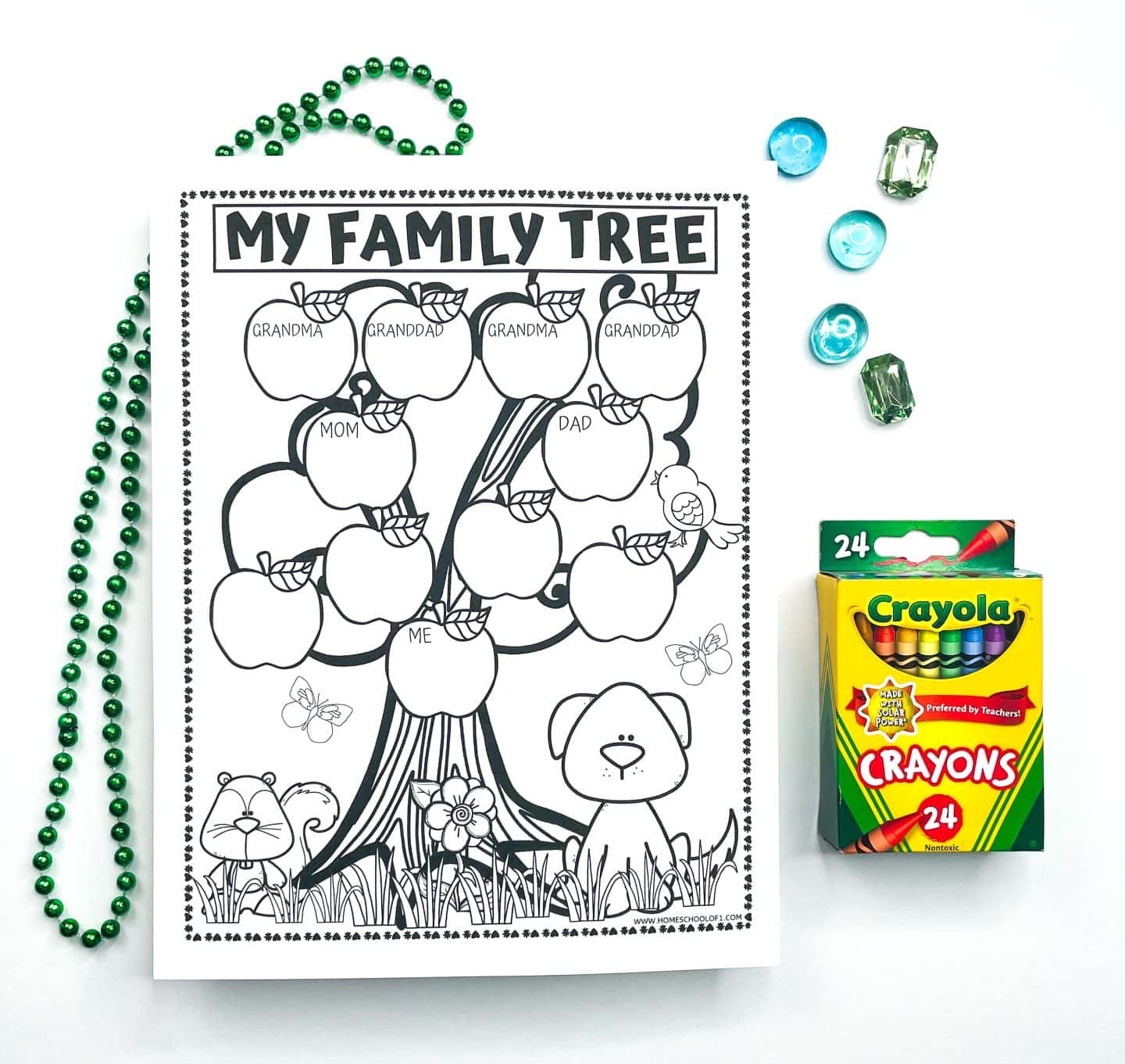 Black-and-white printable “My Family Tree” worksheet for kids, featuring a tree with labeled apples for Grandma, Granddad, Mom, Dad, and Me, plus additional blank apples for extra family members. The bottom of the page is decorated with a dog, squirrel, flower, and butterflies. A box of Crayola crayons sits on the right, with green beads and colorful plastic gems adding a fun craft element.

