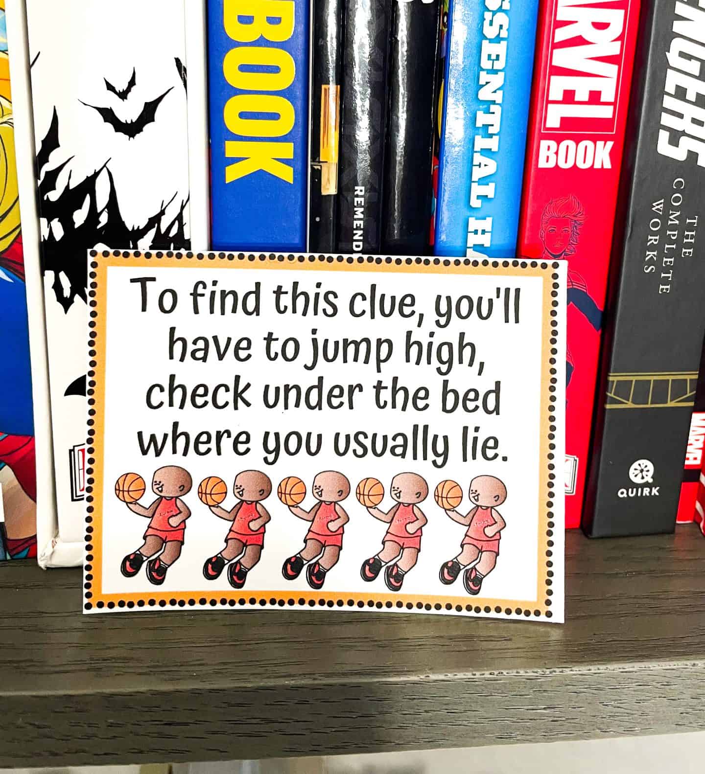 A basketball-themed scavenger hunt clue card displayed on a bookshelf filled with comic and graphic novels, including Marvel and Avengers titles. The card reads: “To find this clue, you’ll have to jump high, check under the bed where you usually lie,” and features a row of cartoon basketball players in red jerseys dribbling basketballs.