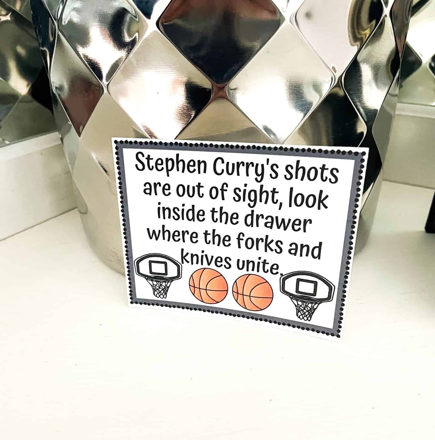 A basketball-themed scavenger hunt clue card placed in front of a shiny, silver diamond-patterned container. The card reads: “Stephen Curry’s shots are out of sight, look inside the drawer where the forks and knives unite,” and is decorated with two basketball hoops and two basketball icons.