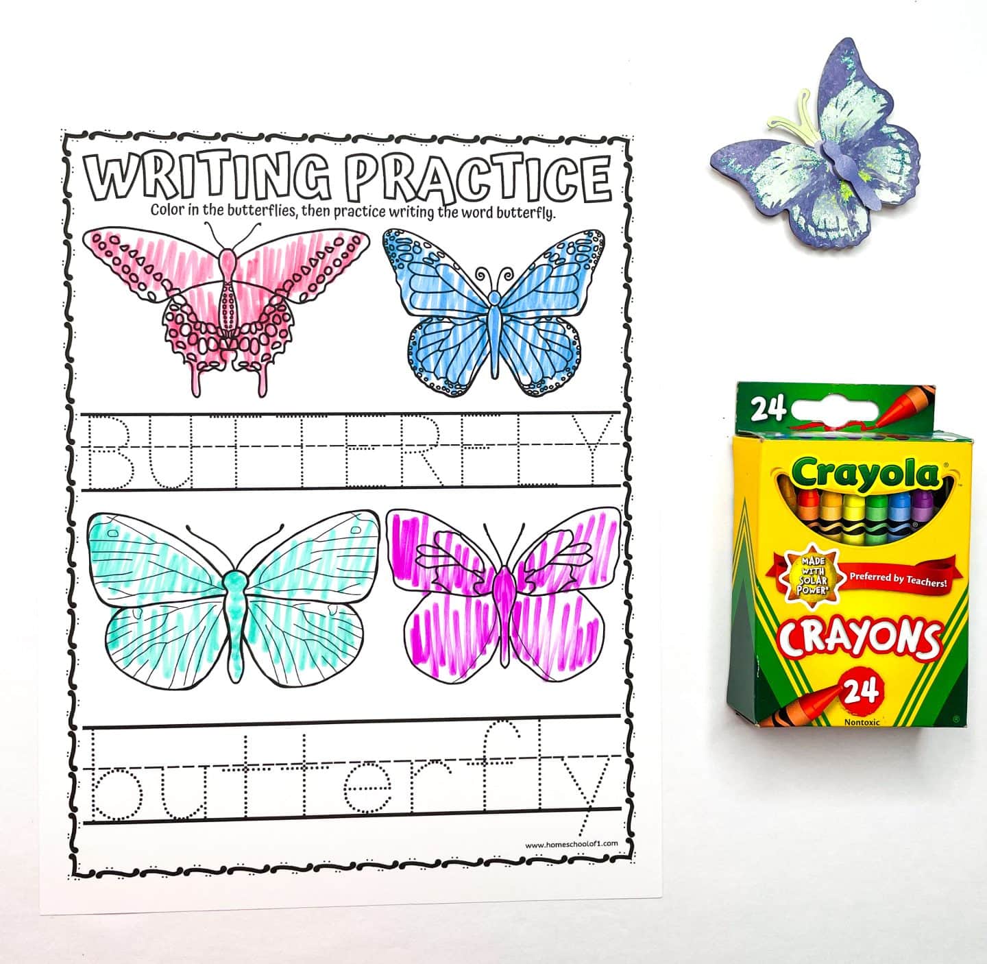 A butterfly writing practice worksheet for preschoolers with four colorful butterflies and two lines for tracing the word "butterfly" in uppercase and lowercase. The butterflies are colored pink, blue, teal, and purple. A 24-pack of Crayola crayons and a decorative purple butterfly are placed beside the worksheet.