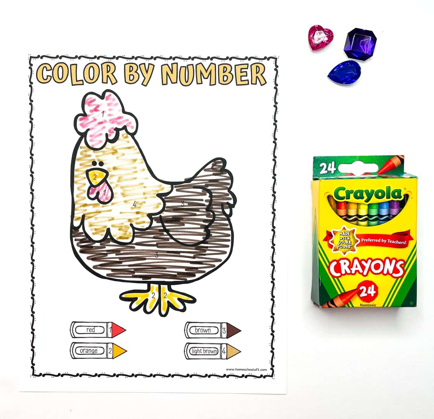 A color-by-number sheet featuring a cartoon chicken with marked sections for red (comb), orange (beak), brown (body), and yellow (feet). Beside it is a box of Crayola crayons and three colored gems.
