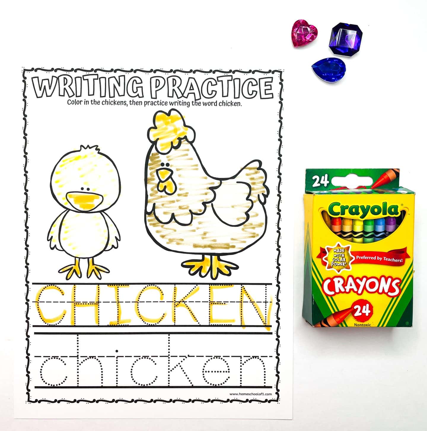 A writing practice worksheet featuring a chick and a hen outlined for coloring, with the word CHICKEN to trace. Beside it is an open 24-pack of Crayola crayons and three plastic jewels.
