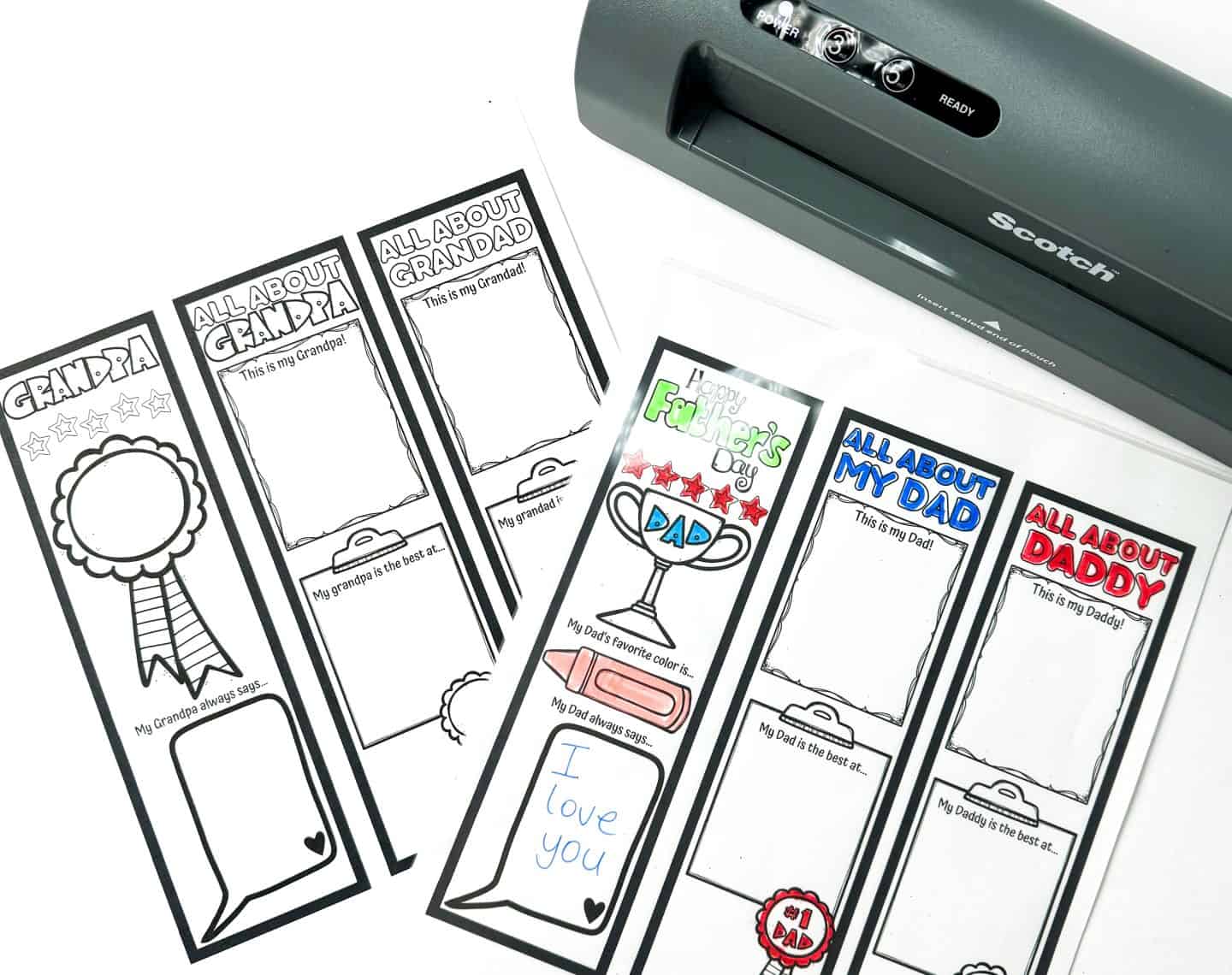 Father’s Day bookmarks being laminated, with some fully colored and filled in. One says “Happy Father’s Day” with red stars, a crayon, and a handwritten message: “I love you.” Other bookmarks include prompts like “This is my Dad” and “My Grandpa always says…” with designs for Dad, Daddy, Grandpa, and Grandad.