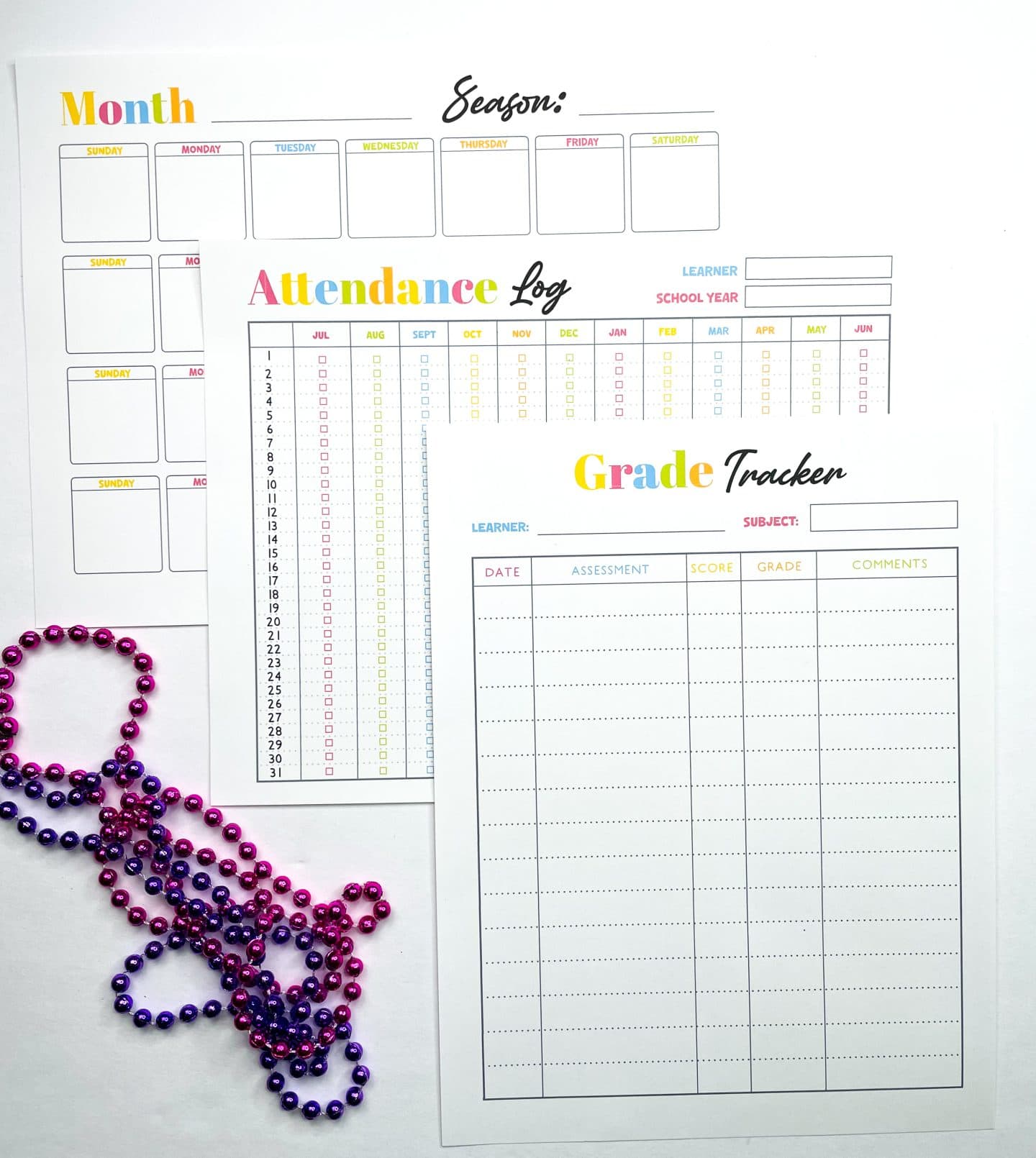 Three colorful homeschool planner printables are laid out: a monthly calendar, an attendance log, and a grade tracker. Each sheet uses bright rainbow-themed headers and clean, lined formatting. Purple and pink plastic bead necklaces are coiled in the lower left for decoration.

