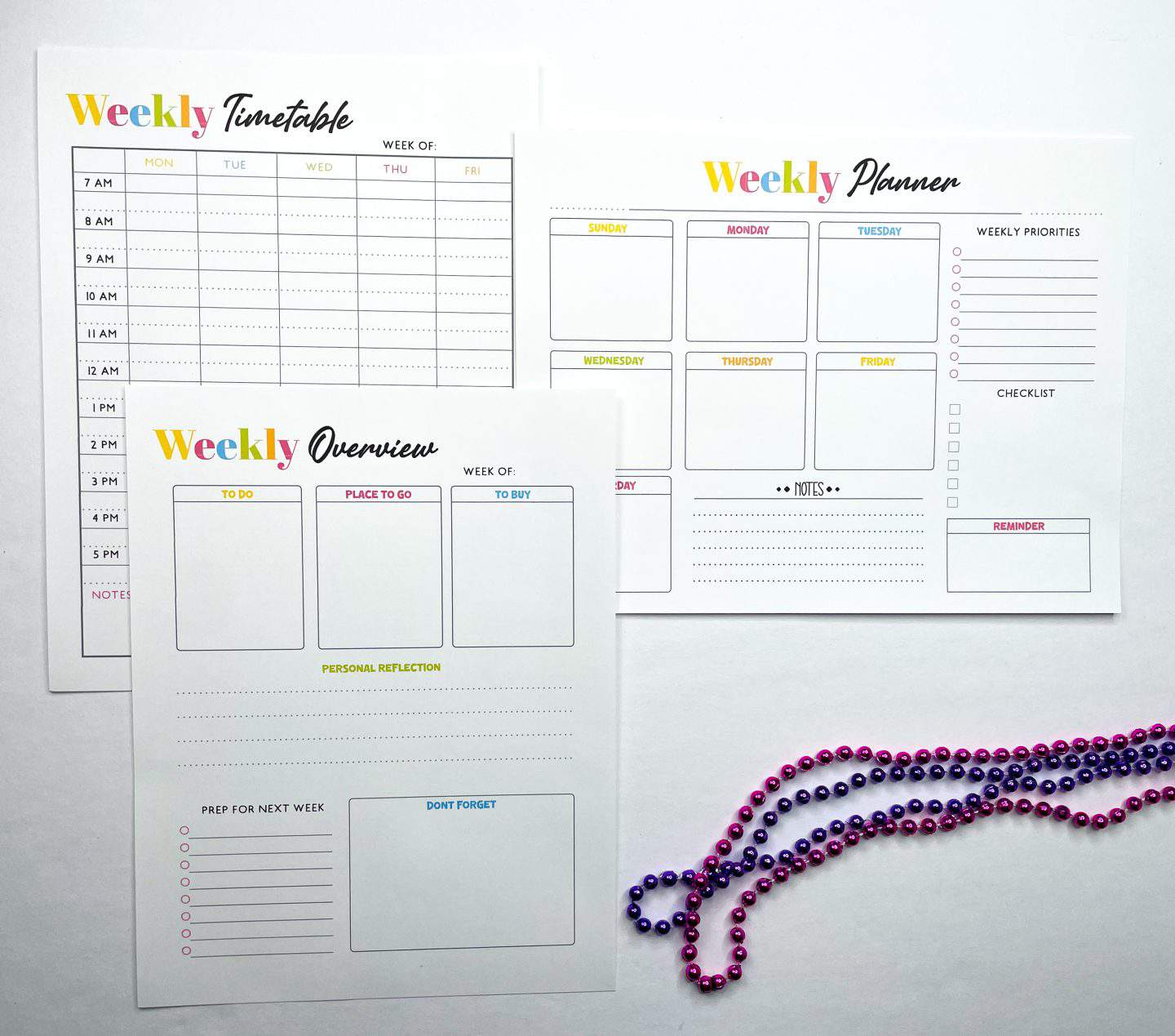 Three printable homeschool planner pages are displayed: a weekly timetable with hourly slots, a weekly planner with daily boxes and priorities, and a weekly overview sheet for tasks, shopping, and reflections. Each page features a cheerful rainbow color scheme and clean, structured design. A strand of purple and pink beads lies along the bottom right corner for decoration.