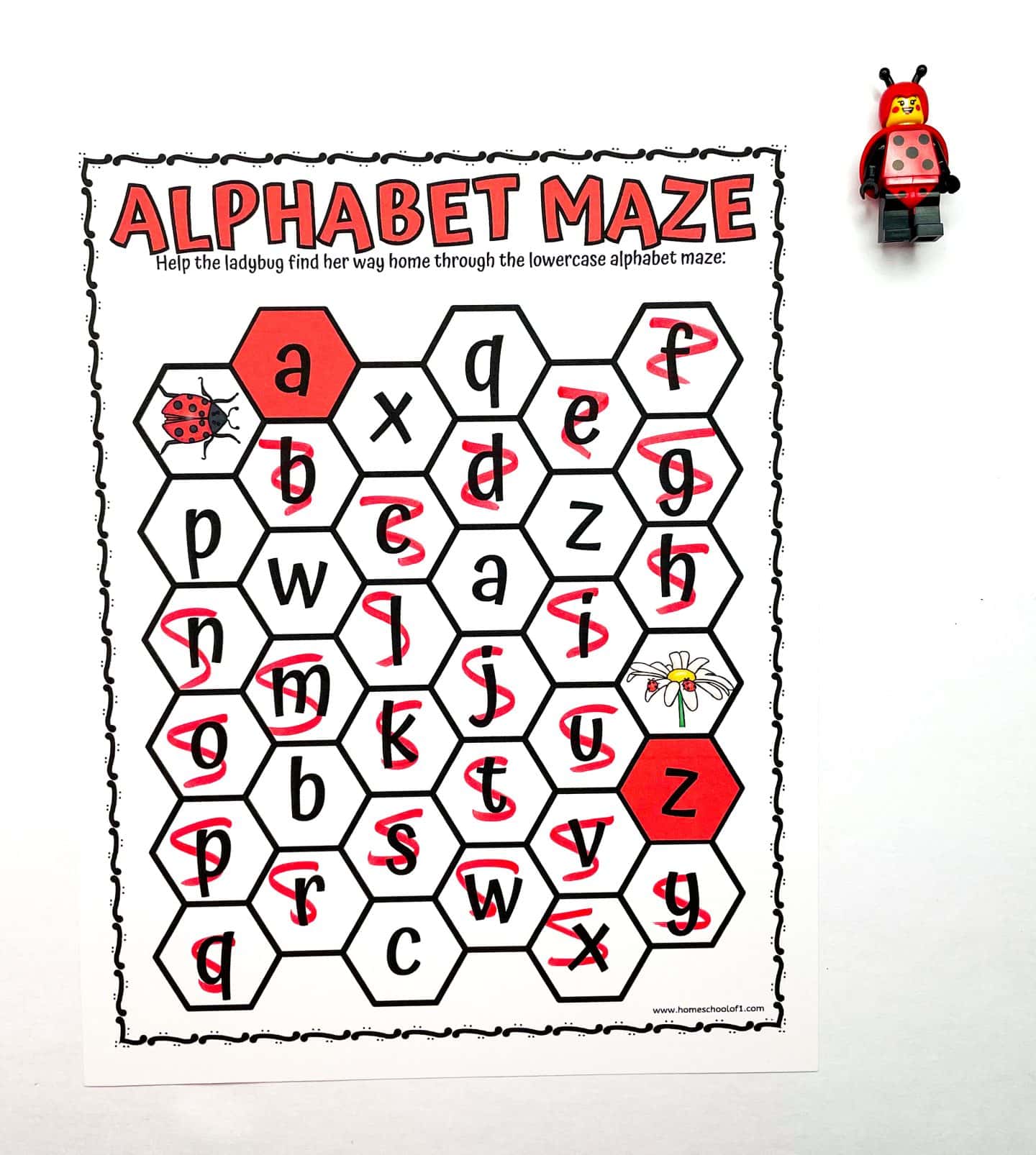 A preschool worksheet titled “Alphabet Maze” features a honeycomb-style grid of lowercase letters. A ladybug icon marks the start at the letter “a” on the left, and a daisy flower marks the end near the letter “z” on the right. A red path is drawn through the correct letter sequence guiding the ladybug home. A LEGO ladybug figurine is placed in the top right corner of the white background.