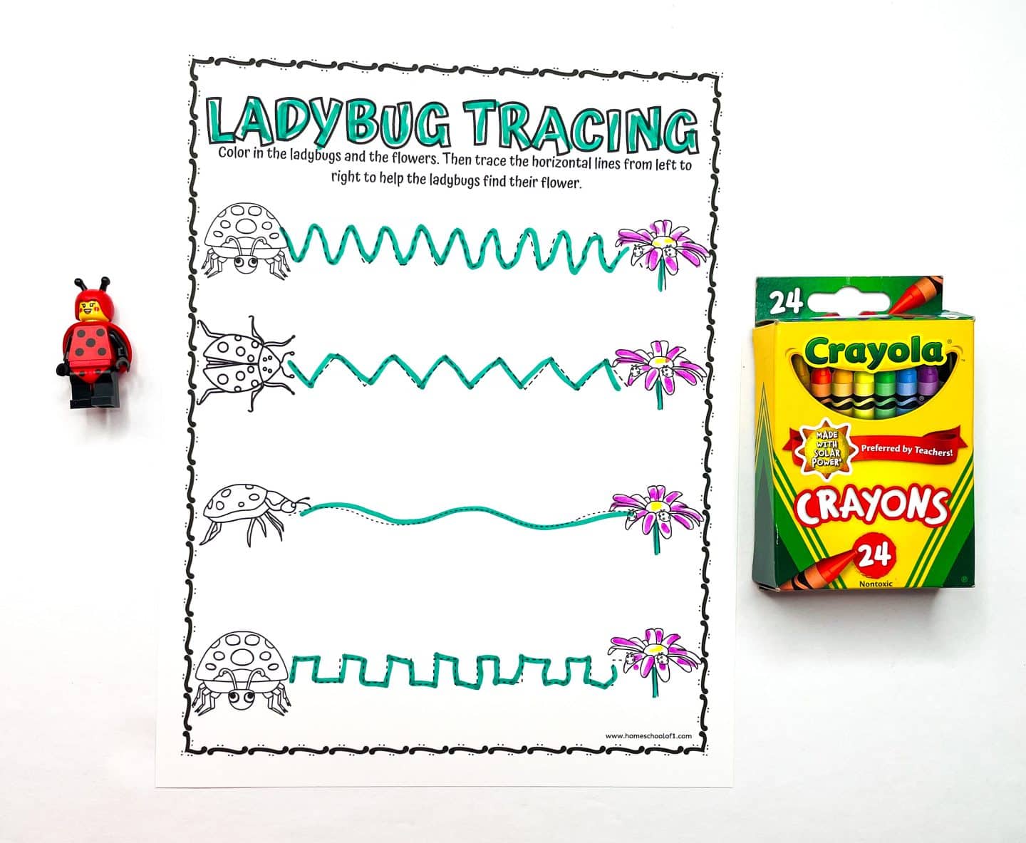 A preschool worksheet titled “Ladybug Tracing” features four horizontal paths with different line patterns for tracing, leading ladybugs to flowers. The paths are outlined in teal, and the flowers at the end of each line are colored pink and purple. A LEGO ladybug figure is on the left, and a box of Crayola 24-count crayons is on the right, all placed on a white background.