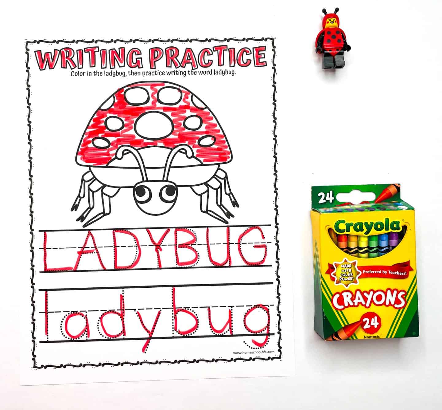 A preschool worksheet titled “Writing Practice” features a large cartoon ladybug partially colored red and the word “ladybug” written in uppercase and lowercase letters with red dotted tracing lines. A box of Crayola 24-count crayons and a small LEGO ladybug figure are placed next to the worksheet on a white background.
