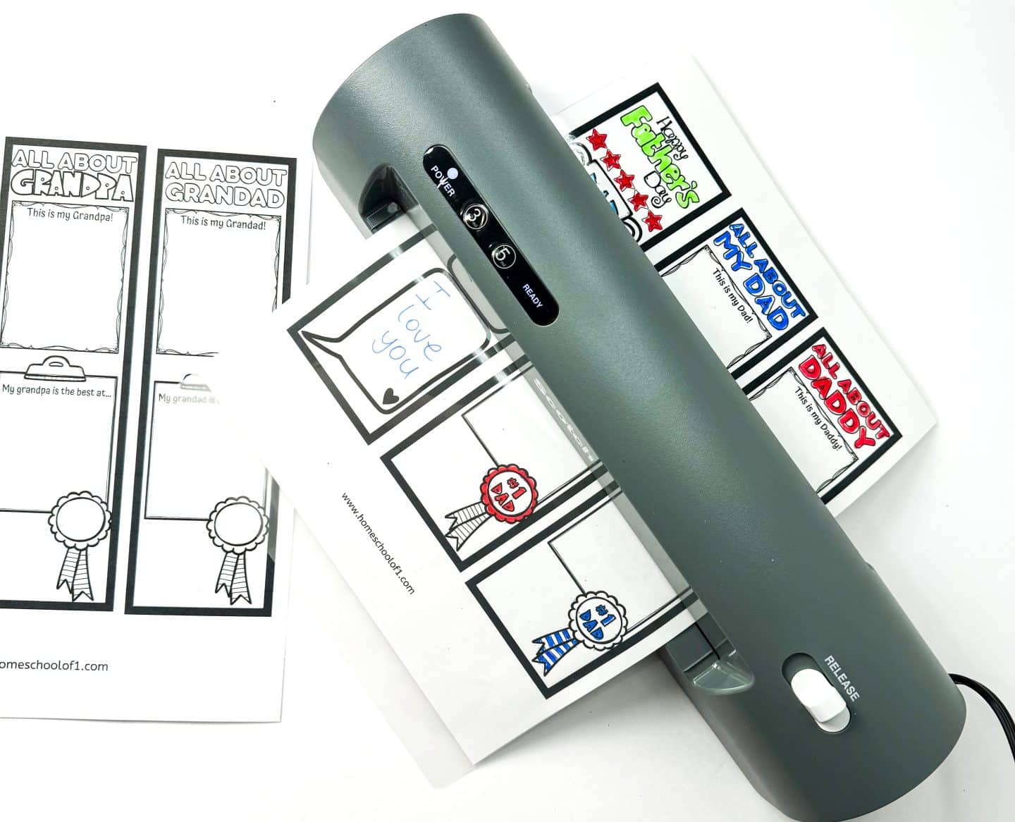 A laminator sealing a sheet of colorful Father's Day bookmarks, including one with the message “I love you” written in blue. The designs read “Happy Father’s Day,” “All About My Dad,” and “All About Daddy,” while uncolored versions for “Grandpa” and “Grandad” sit nearby, ready to be filled out and customized.

