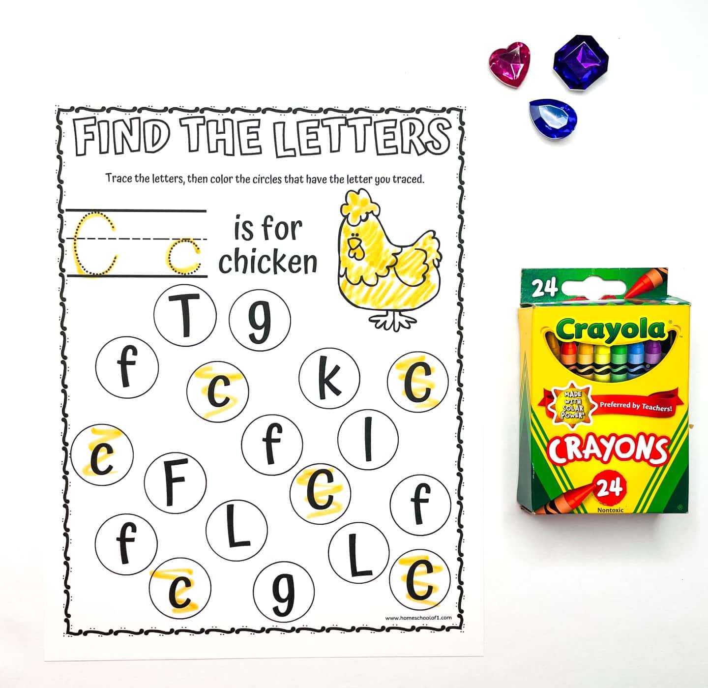 A worksheet titled Find the Letters featuring a chicken illustration. It says C is for chicken and includes a tracing activity with letter C circles. Three colorful gems and a box of Crayola crayons are beside the sheet.
