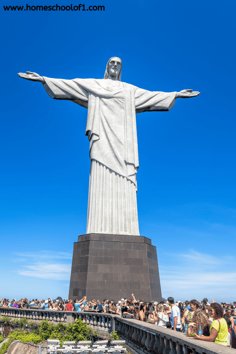 Tourists gather at the base of the towering Christ the Redeemer statue in Rio de Janeiro, Brazil, under a clear blue sky.