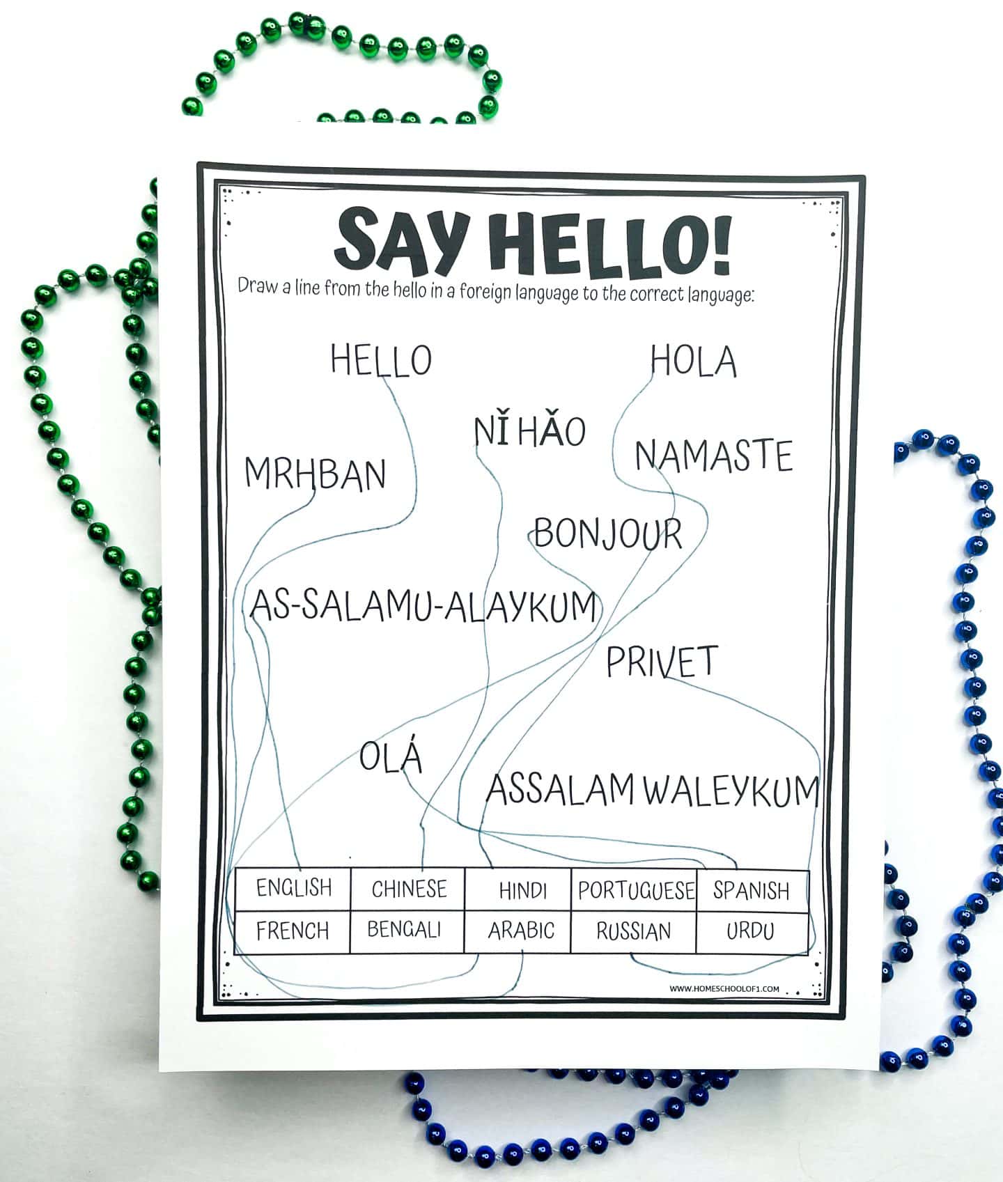 A printable worksheet titled "Say Hello!" featuring a matching activity. It asks students to draw lines connecting various greetings in different languages (e.g., "Hola," "Bonjour," "Namaste") to the correct language labels at the bottom, such as English, Spanish, French, Arabic, and more. Curved lines show completed matches. Green and blue beaded necklaces frame the worksheet.