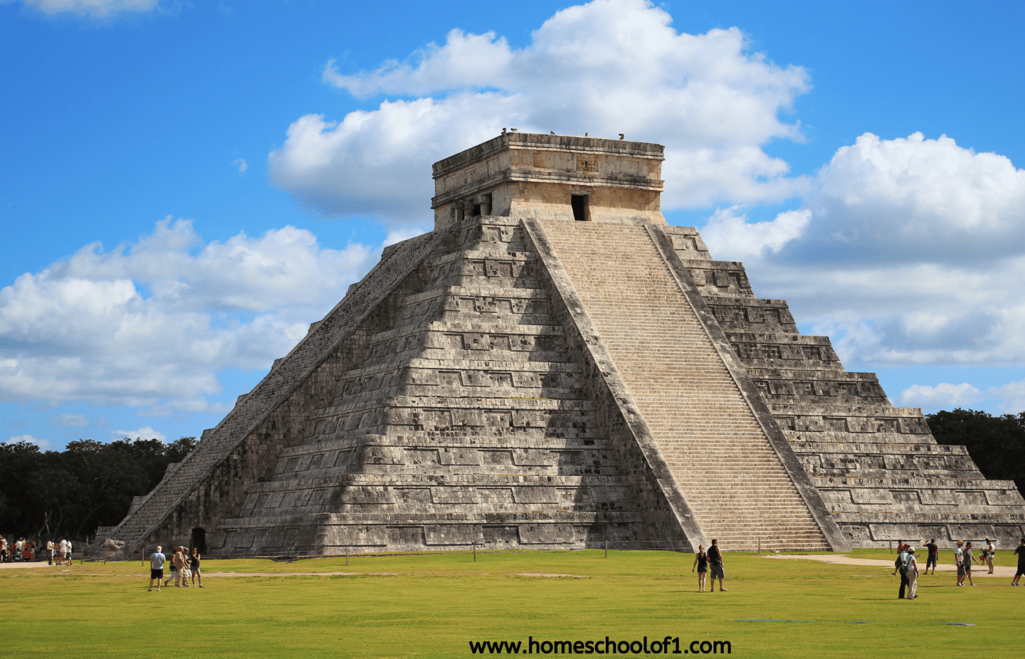 Photo of the Chichen Itza pyramid under a partly cloudy blue sky, with tourists walking around the grassy area in front.
