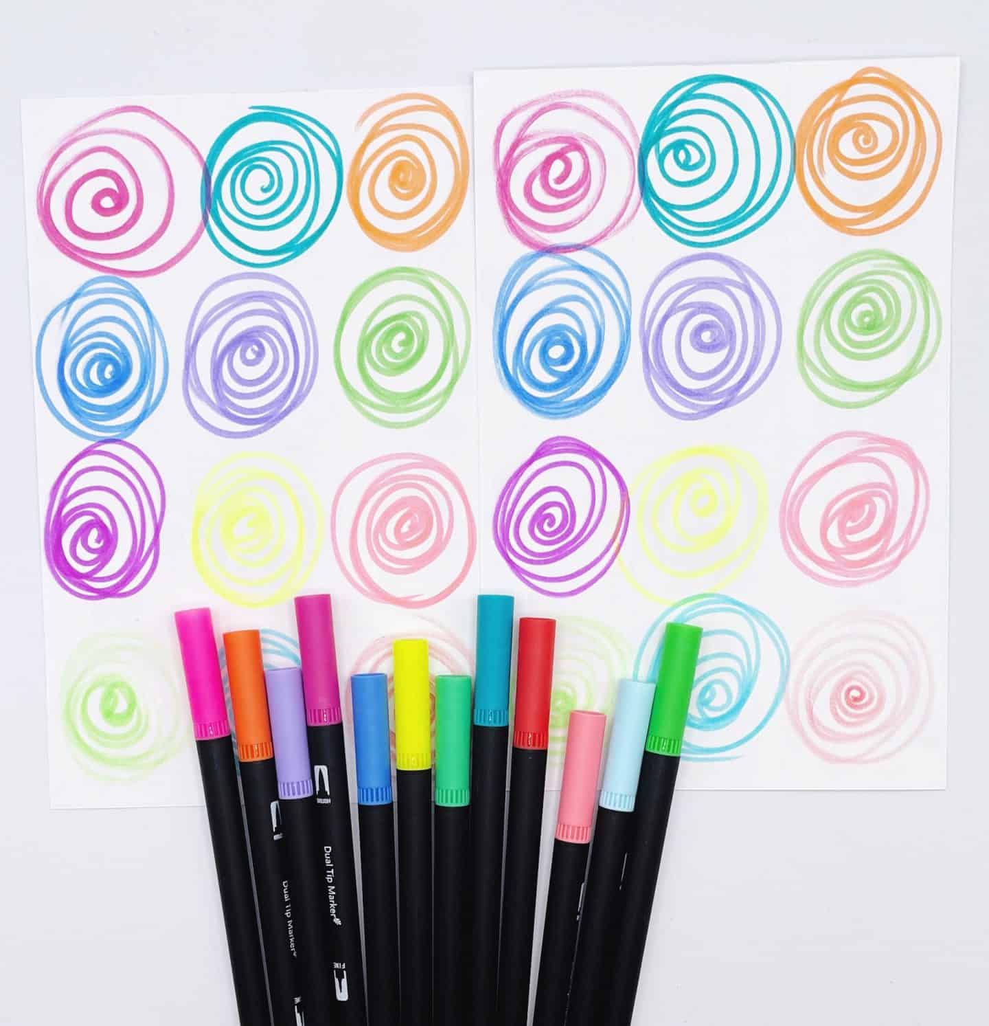 Two sheets of paper with colorful hand-drawn spiral patterns in matching pairs, designed for a memory game. Various markers in coordinating colors—pink, orange, green, blue, purple, and yellow—are laid out at the bottom, showing the tools used to create the designs.