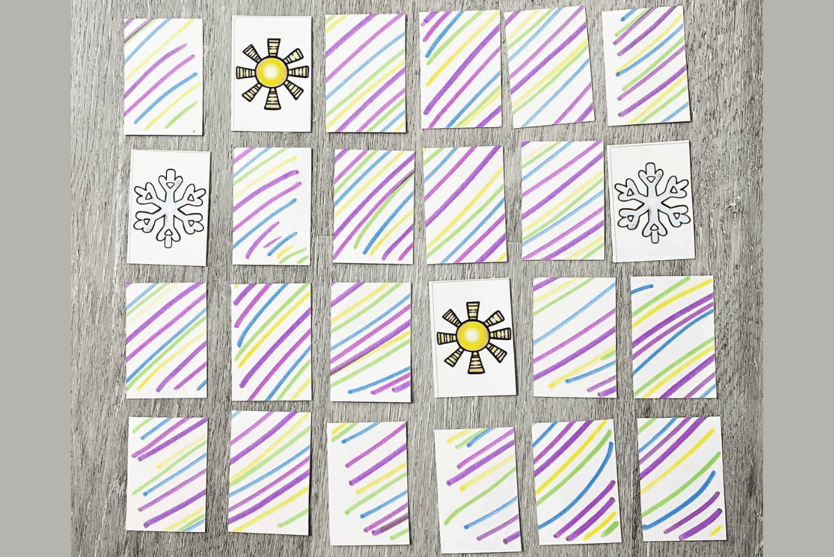 Printable weather memory matching game for kids with cards laid out in a grid on a wooden surface. Most cards show colorful diagonal stripes on the back, while a few are flipped to reveal weather icons like a sun and a snowflake.