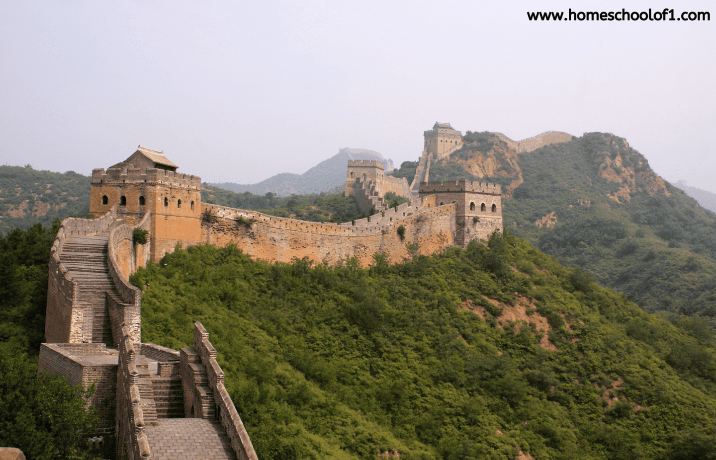 Photograph of the Great Wall of China winding over lush green hills, showcasing its ancient stone architecture, watchtowers, and impressive scale under a hazy sky. 