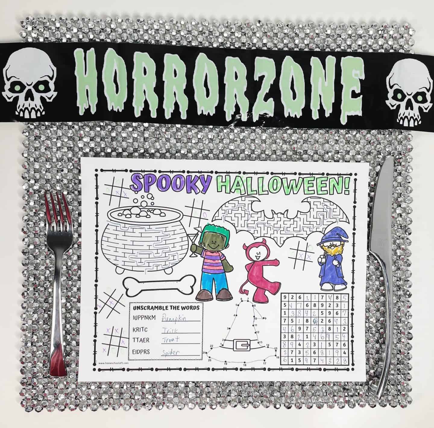 A festive Halloween kids’ placemat featuring activities like mazes, word scramble, and connect-the-dots is placed on a glittery silver charger plate with a fork and knife. The placemat is partially colored and titled “Spooky Halloween!” in bold purple and green text. Characters in monster, devil, and wizard costumes are featured. The setup includes a black "Horror Zone" banner with green letters and skull graphics across the top.