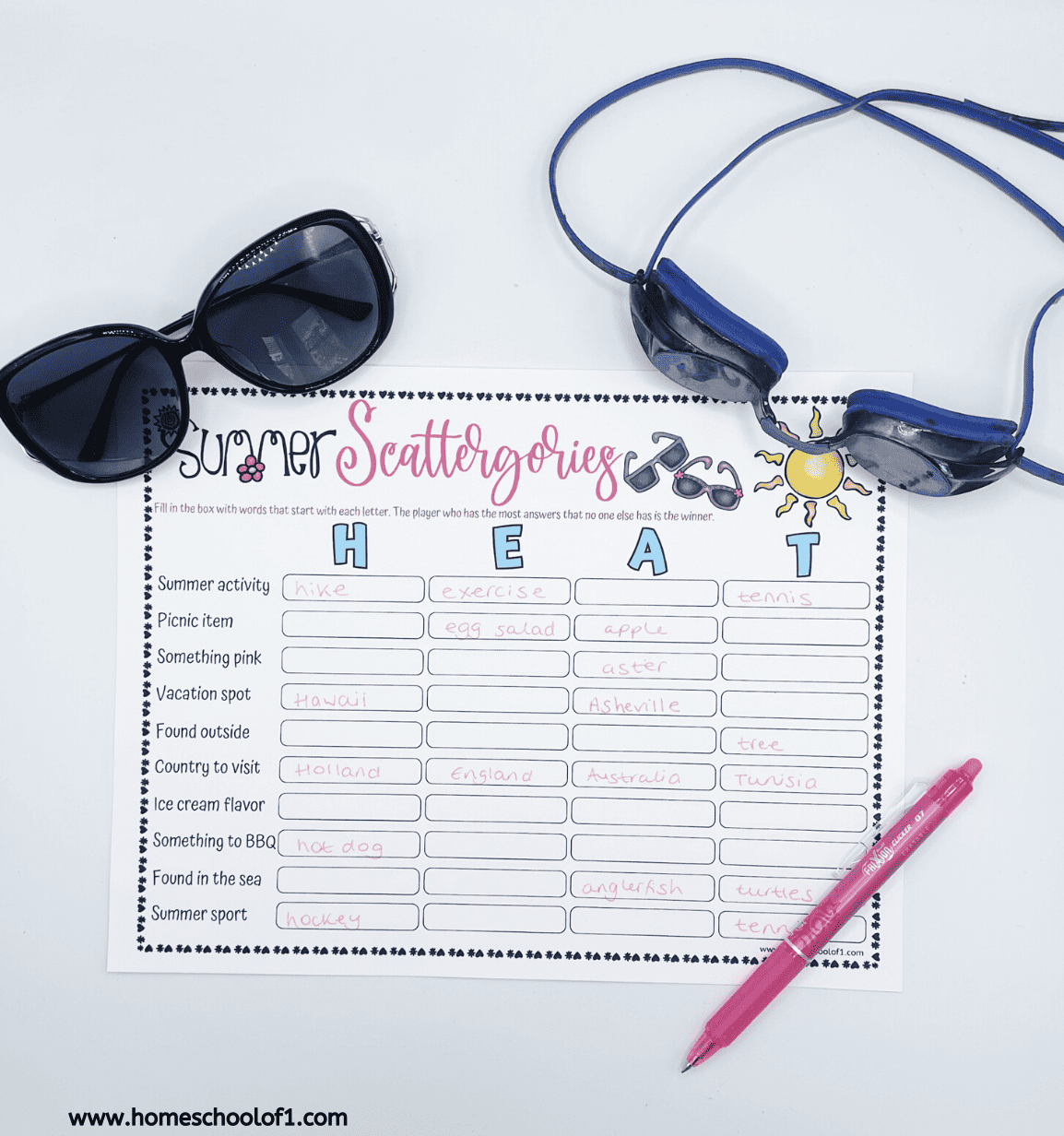 A printed "Summer Scattergories" game sheet is filled out with answers for the word "HEAT" under categories like summer activity, picnic item, and vacation spot. Items include "hike," "egg salad," "Hawaii," "apple," and "tennis." The sheet is surrounded by black sunglasses, blue swim goggles, and a pink pen, all on a white surface. 