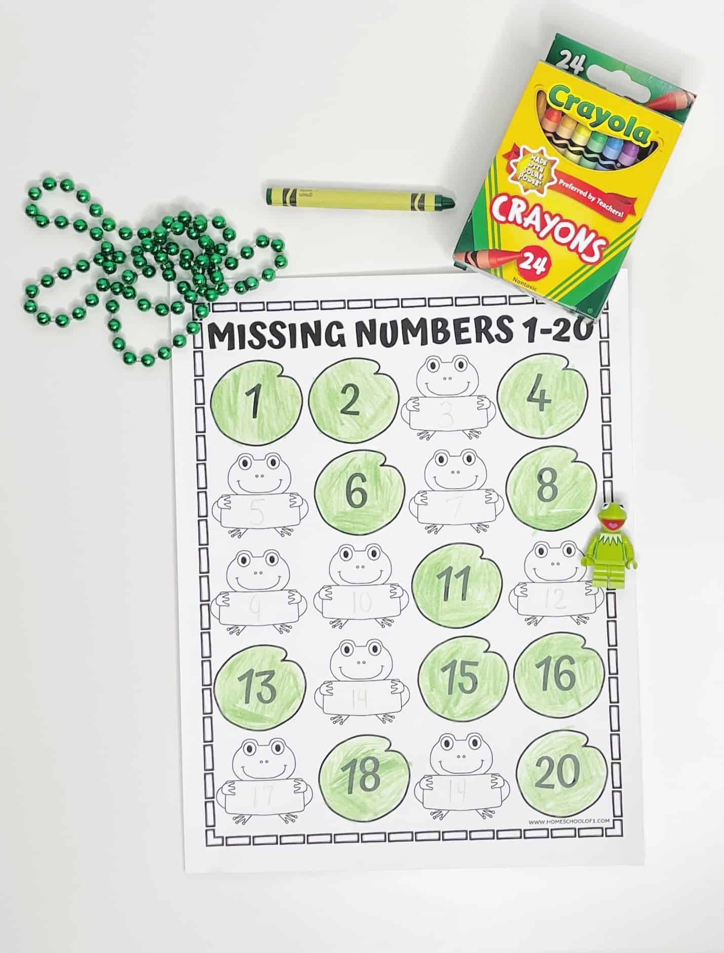  A “Missing Numbers 1–20” worksheet with a frog and lily pad theme, partially filled in with green crayon. Each lily pad contains a number, and frogs are holding spaces for the missing numbers. The sheet is set on a white surface with a green beaded necklace, a yellow-green crayon, a box of Crayola crayons, and a small Kermit the Frog LEGO figure nearby.